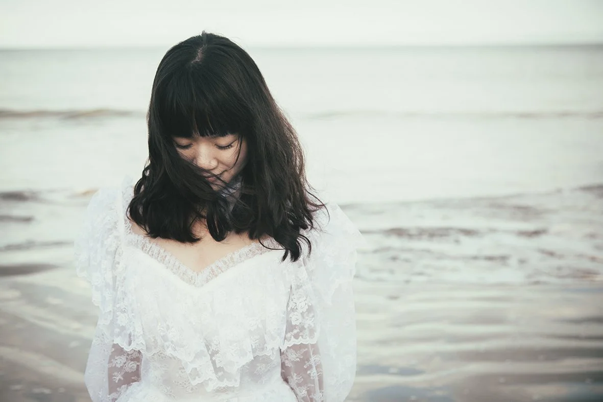 A woman with black hair wearing a white lace dress standing at the beach with gentle waves in the background