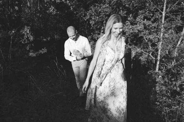 A woman in a floral dress walking on a wooded trail, smiling, with a man in the background looking at his phone.