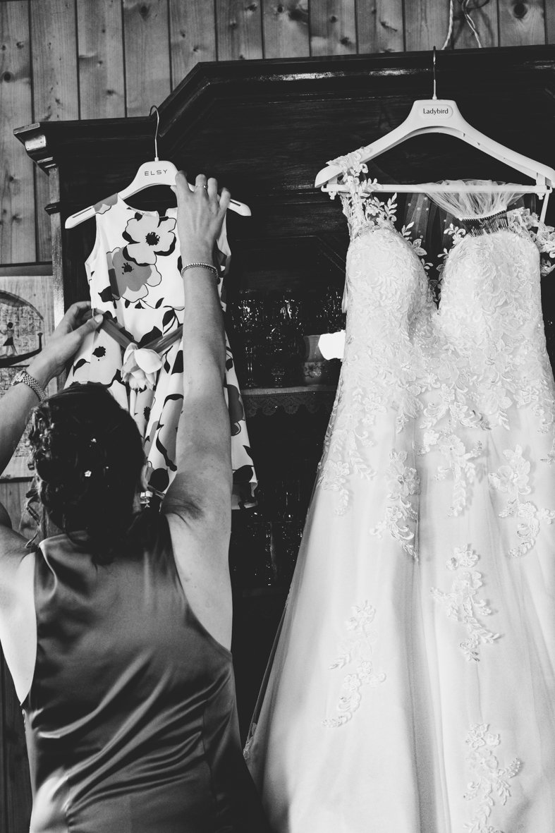 A woman with hair decorated with flowers is holding a floral dress on a hanger. Next to her, a wedding gown with lace and floral embroidery is hanging on a different hanger.