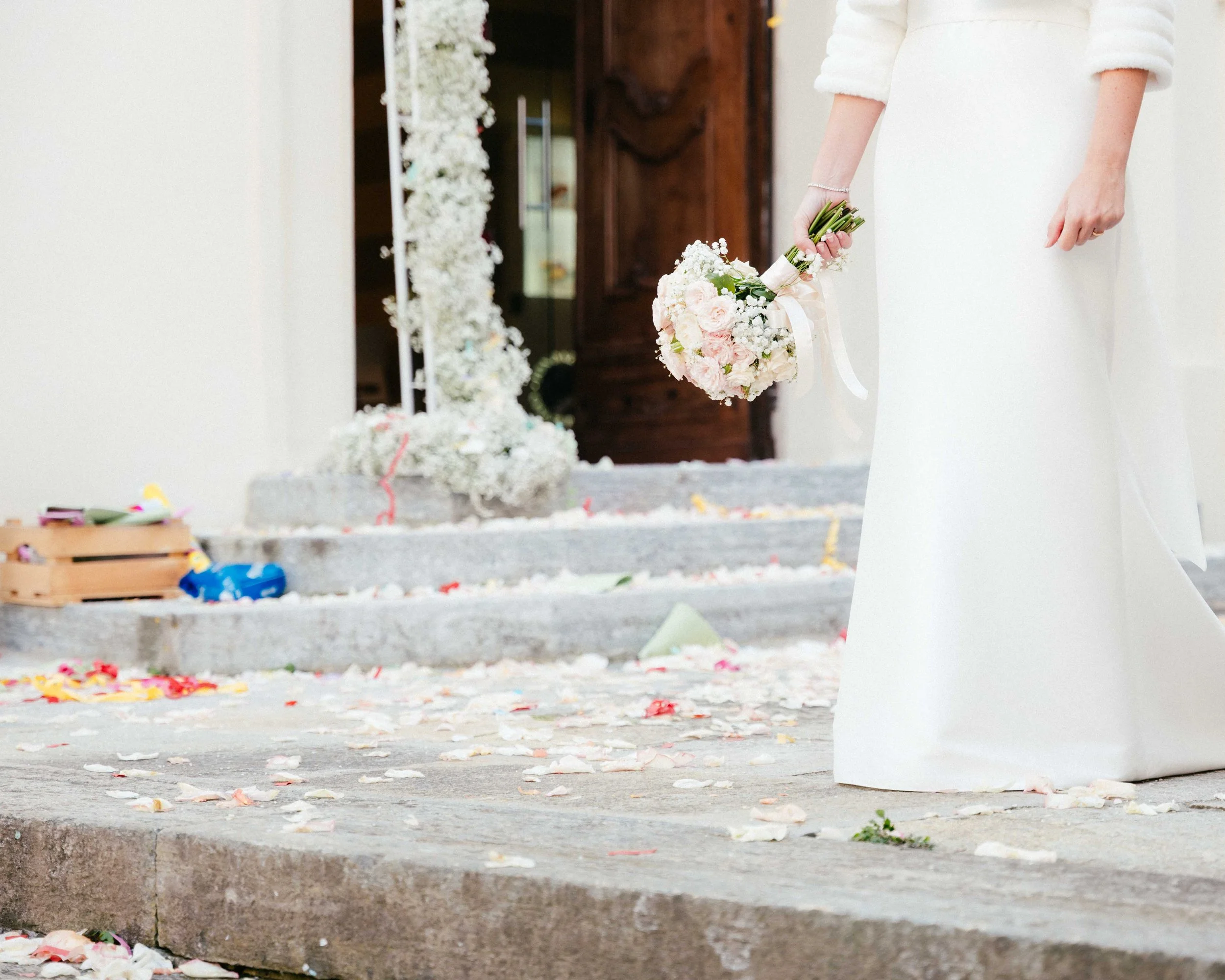 A person in a white dress holding a bouquet of white and pink flowers, standing near a decorated area that appears to be for a wedding or celebration, with scattered flower petals on the ground.