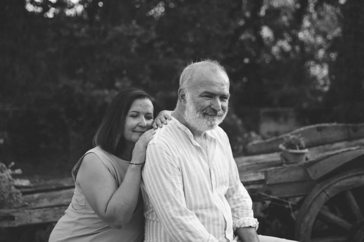 A woman with dark hair embracing a smiling older man with a beard, outdoors near a wooden wagon and trees.