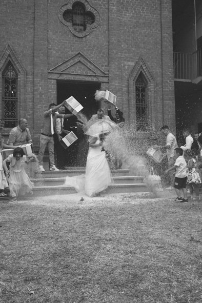 A woman in a wedding dress holding an umbrella, standing outside a church, is splashed with water while several children and adults throw books and water at her.