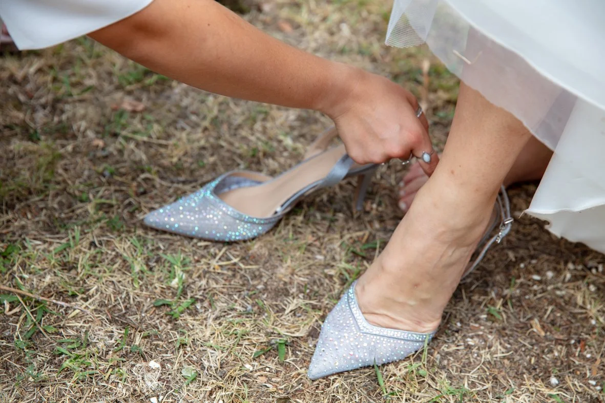 A person is putting on a sparkly high heel shoe as part of a wedding dress, outdoors on grass.