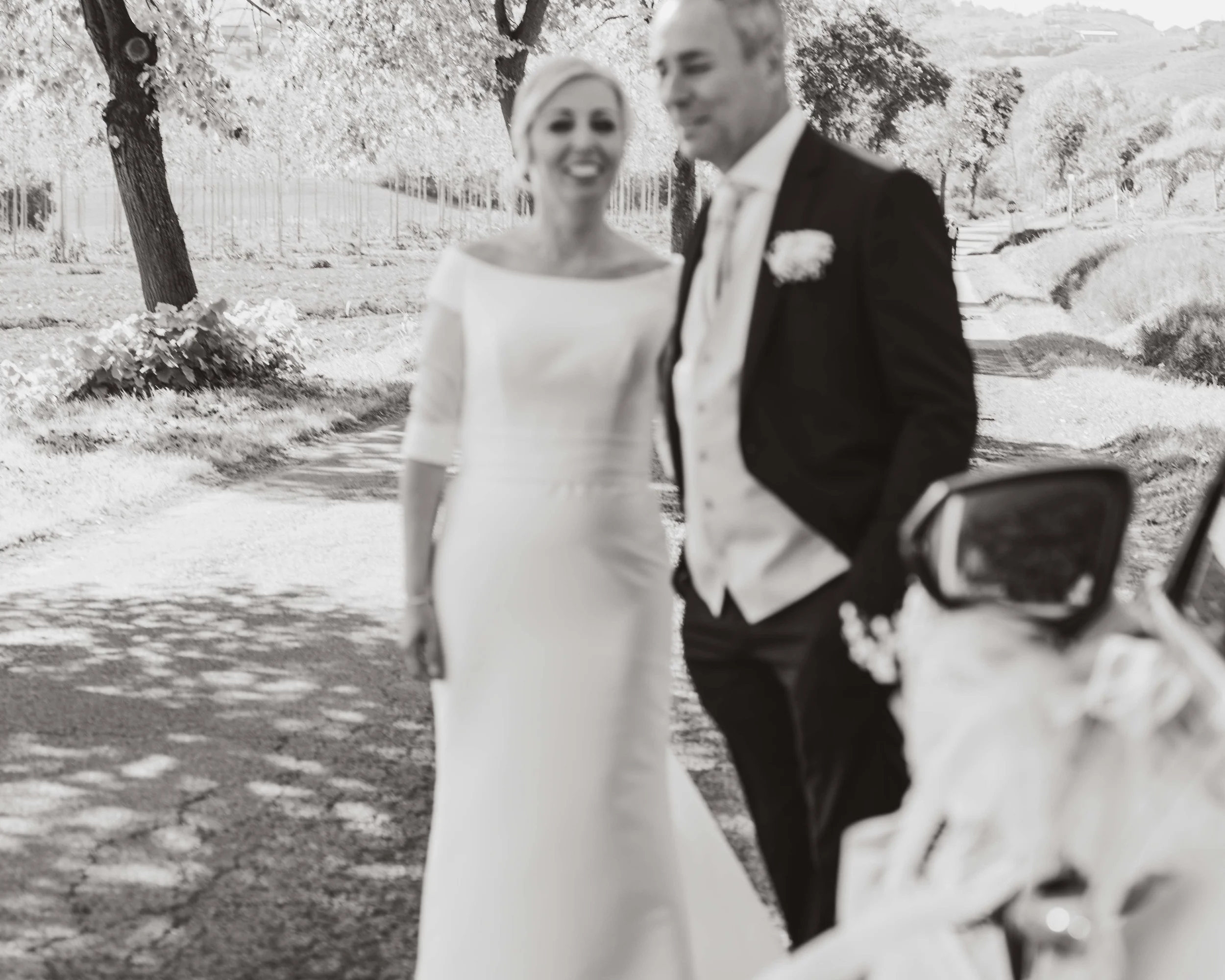 A black-and-white photo of a bride and groom walking outdoors, smiling, with trees and a pathway in the background.