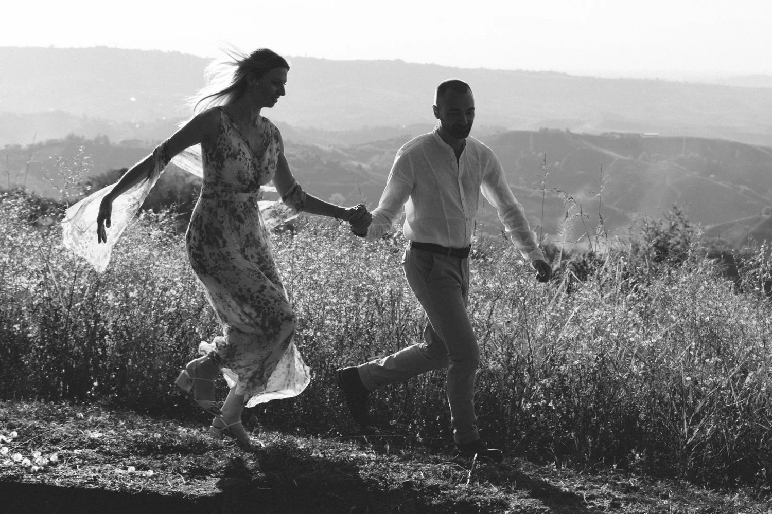 A man and woman running hand in hand through a grassy field on a hillside during sunset, black and white photo.
