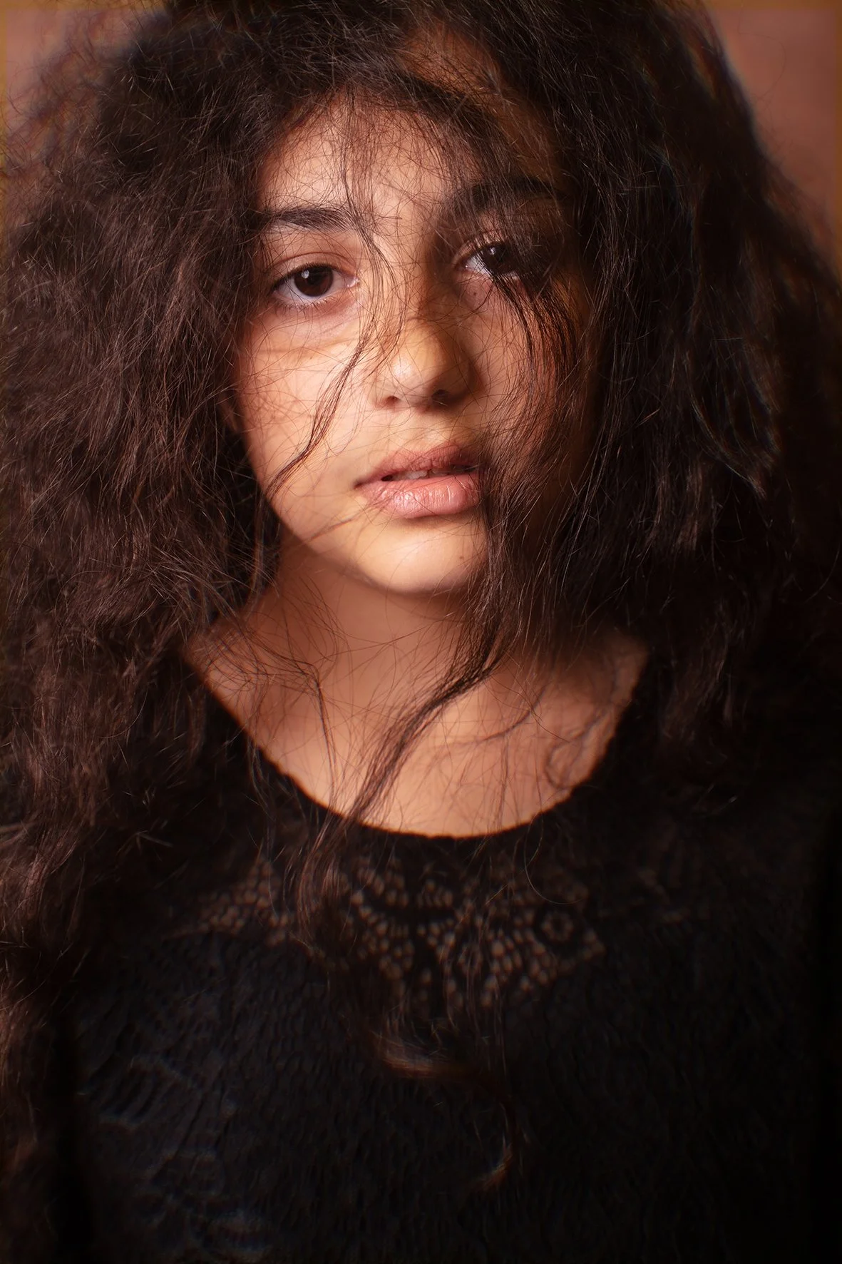Ragazza con capelli ricci e spessi, che indossa un top nero con dettagli in pizzo, guarda verso la fotocamera con un'espressione sfumata.