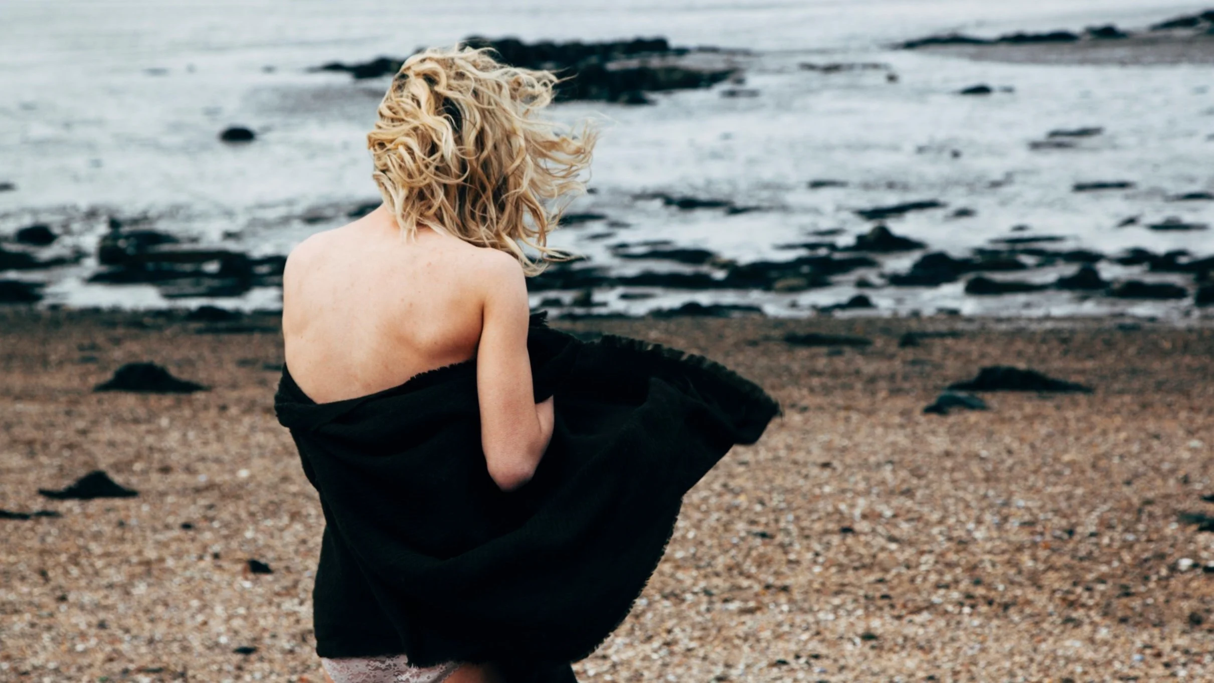Donna nuda seduta sulla spiaggia con un cappotto nero, rivolta verso il mare.