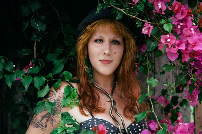 Una donna con capelli ricci castani, indossa un cappello nero, una collana e un vestito a pois, circondata da foglie verdi e fiori rosa