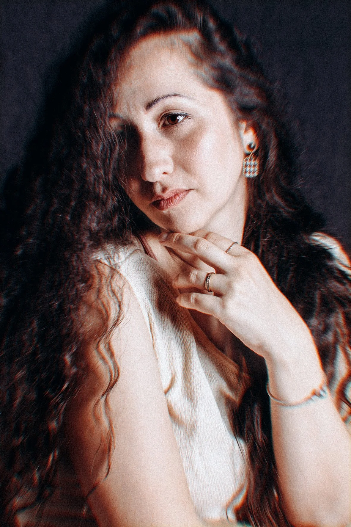  Ritratto di una donna con capelli ricci e lunghi, che indossa orecchini a cerchio e anelli, con una camicia bianca e sfondo scuro.