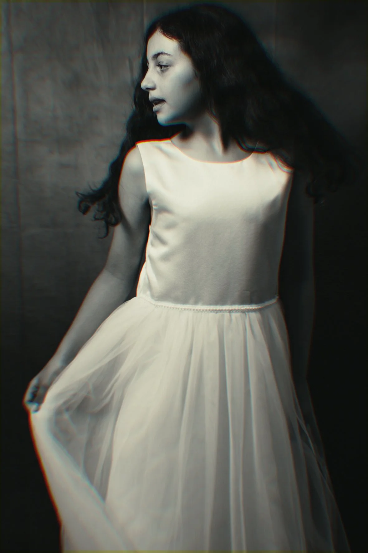 Donna con capelli ricci neri, vestita con un abito bianco, posa con uno sguardo di profilo contro uno sfondo scuro.