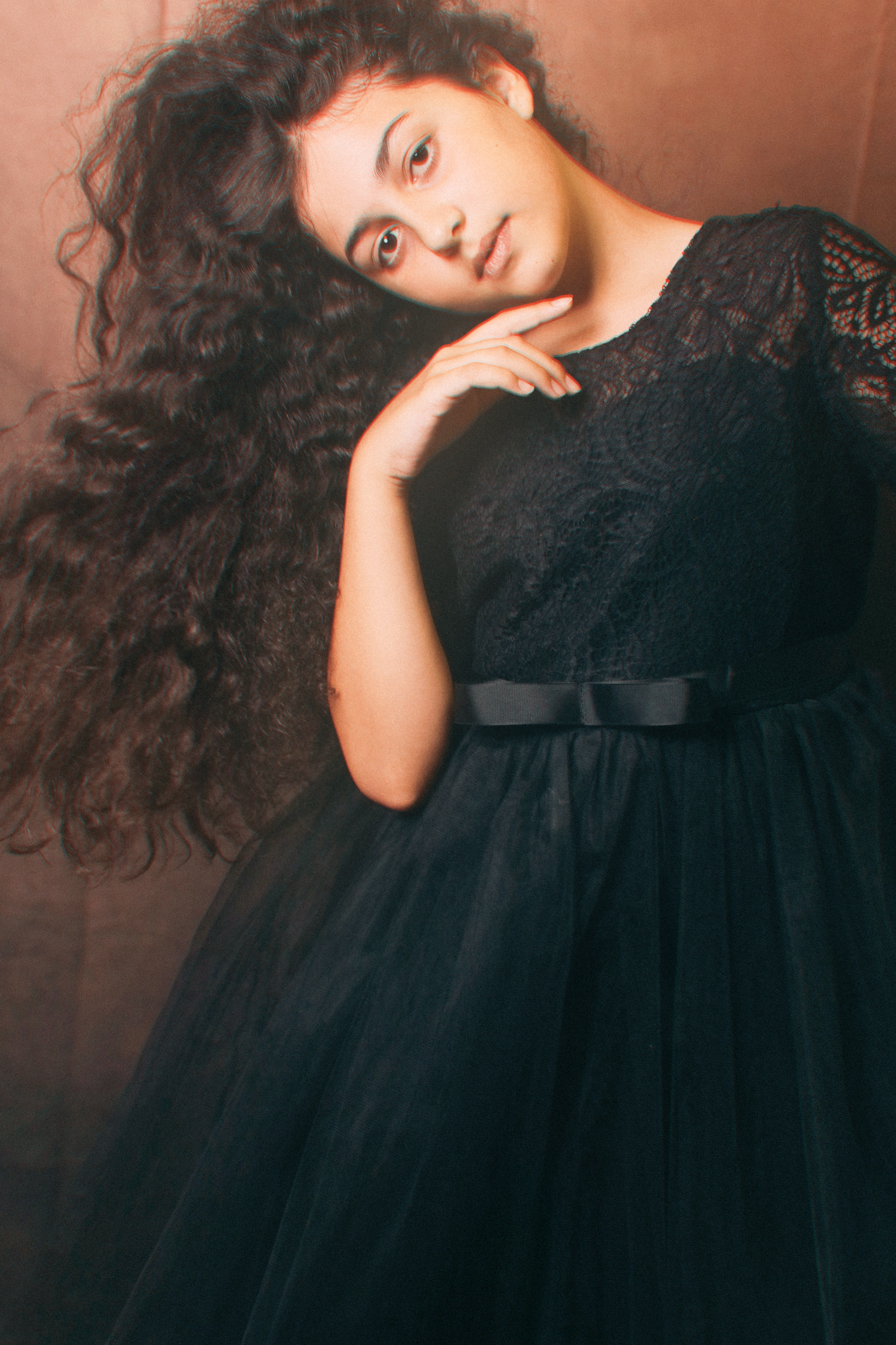 Ritratto di una donna con capelli ricci lunghi e neri, indossa un vestito nero in pizzo e una cintura nera in vita, posa con la testa leggermente inclinata e una mano vicino al viso.