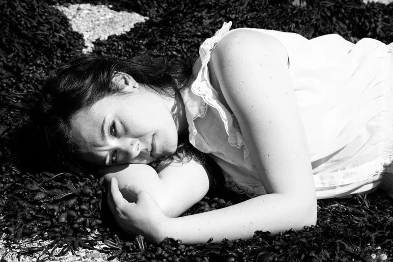 Una donna stesa su un prato con fiori, in abbigliamento bianco, che guarda verso la fotocamera in modo tranquillo.