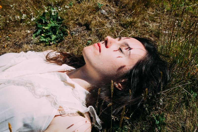 Una giovane donna con capelli scuri e abbigliamento bianco giace a terra tra erba e piante, con gli occhi chiusi e un'espressione serena.