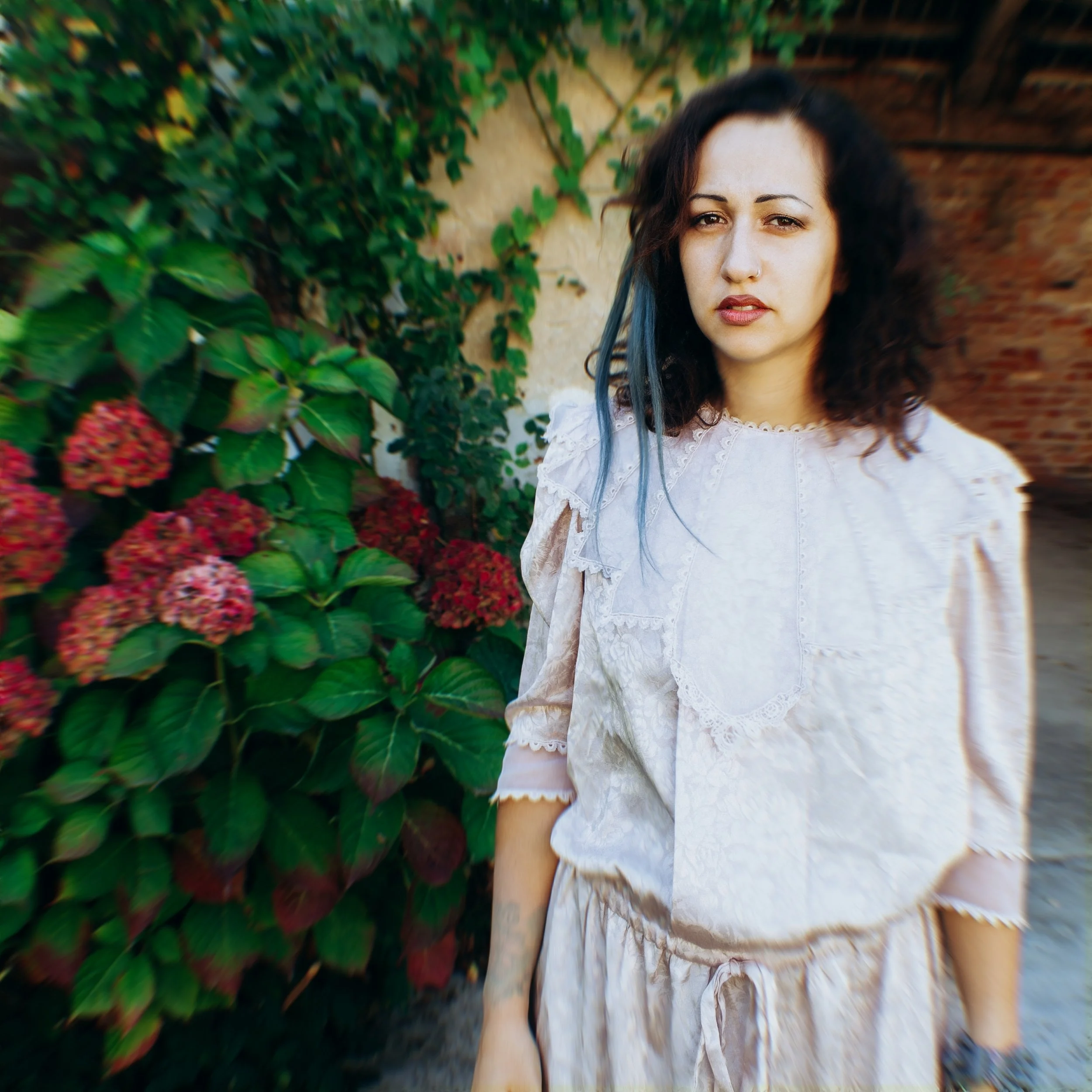 Ragazza con capelli ricci neri e una fascia blu, in abito bianco con dettagli a pizzo, davanti a un cespuglio di ortensie rosse.
