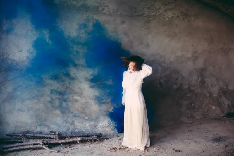 Una donna in abito bianco che sorride, con i capelli che sventolano, in una stanza con pareti in cemento e un pavimento di terra, con un filo di fumo blu nell'aria.