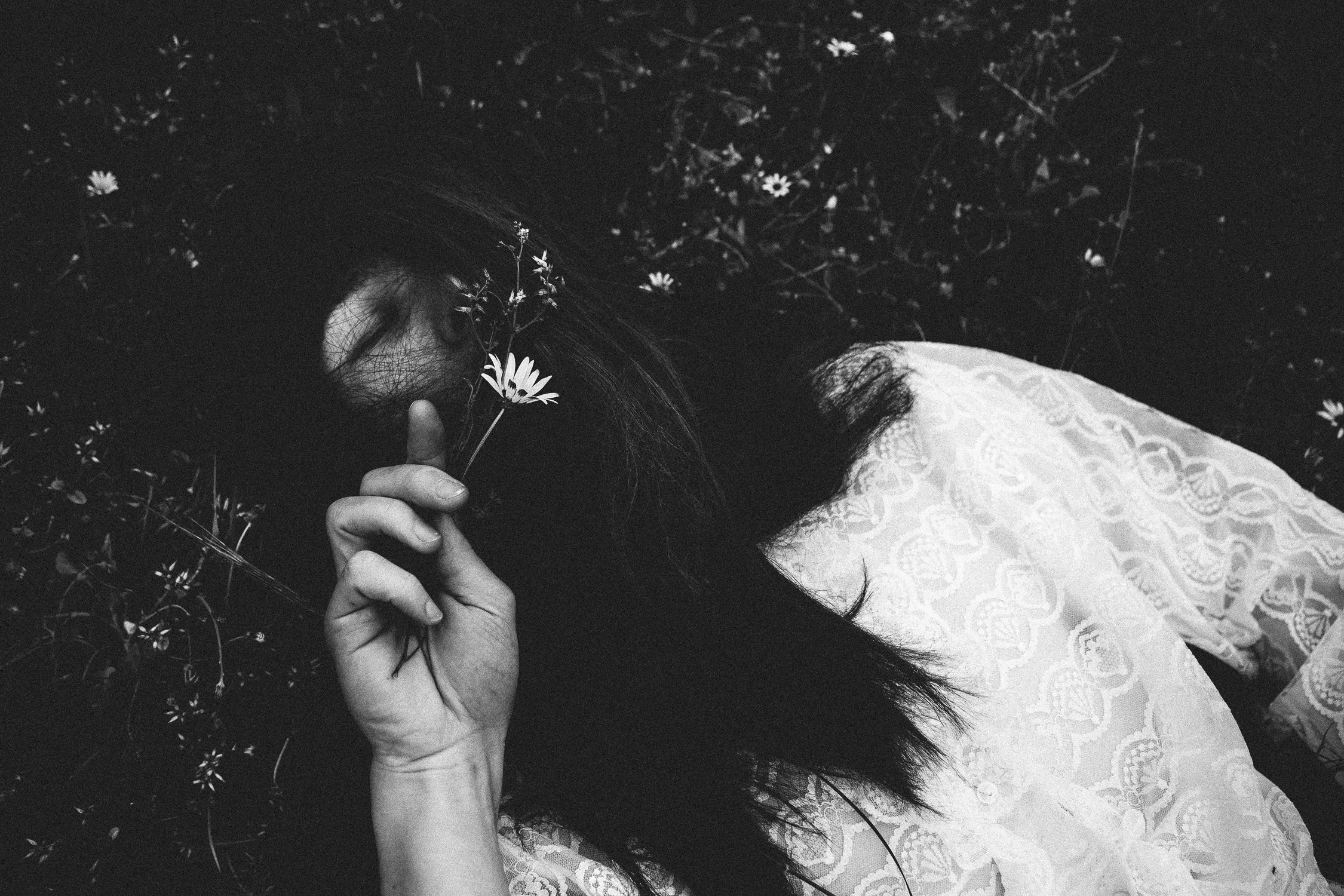 Donna sdraiata tra l'erba con fiori, coperta con un abito bianco ricamato, che tiene un fiore davanti al viso, in un contesto naturale.