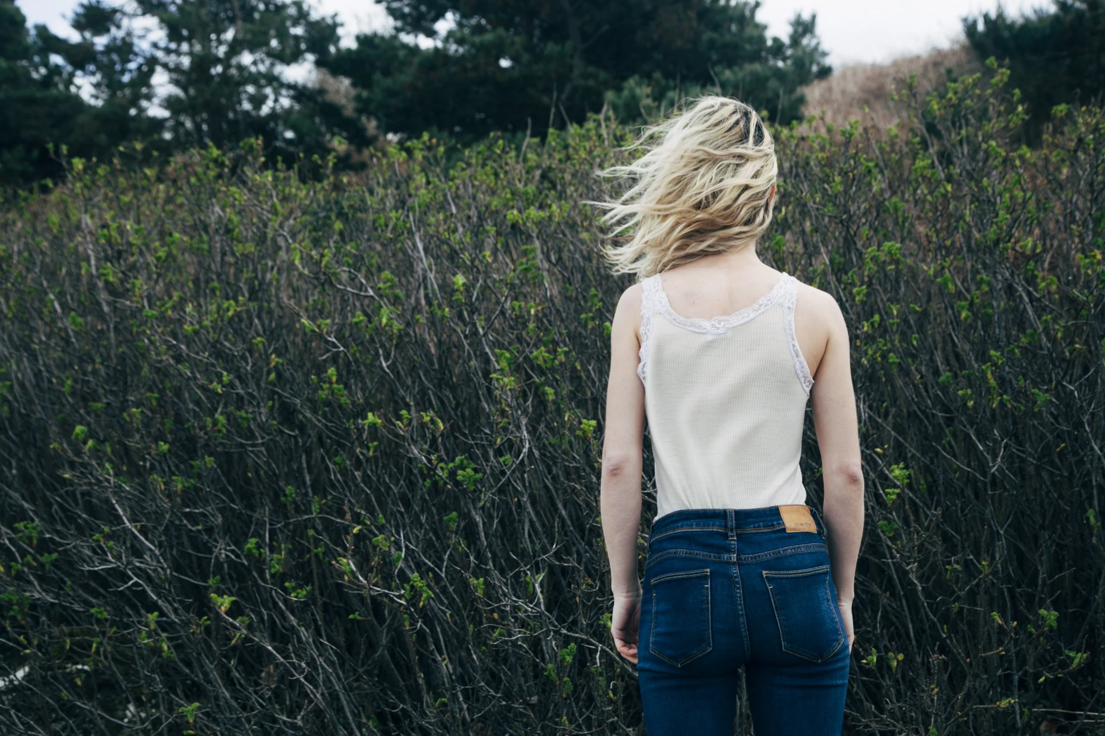 una donna con capelli biondi corti, indossa un top bianco e jeans blu scuri, vista di schiena, davanti a una siepe di rami spinosi con foglie verdi