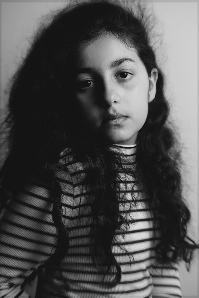 Ragazza giovane con lunghi capelli ricci, vestita con maglia a righe, posa di fronte a uno sfondo neutro.