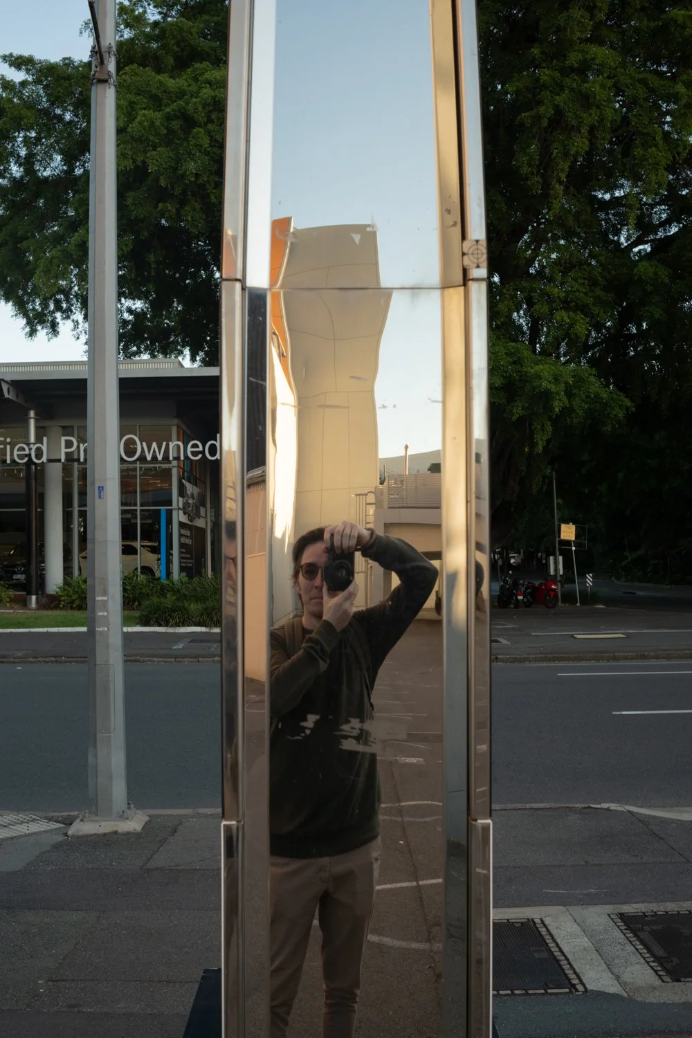 Reflection of a person taking a photo in a tall, vertical mirror outside on a sidewalk, with trees and a building in the background.