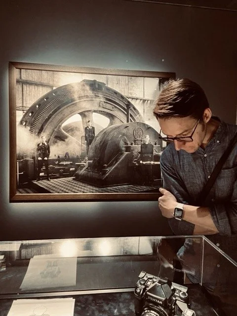 A man looking down at photography equipment displayed on a glass table, with a large black and white photograph of a vintage industrial machine on the wall behind him.
