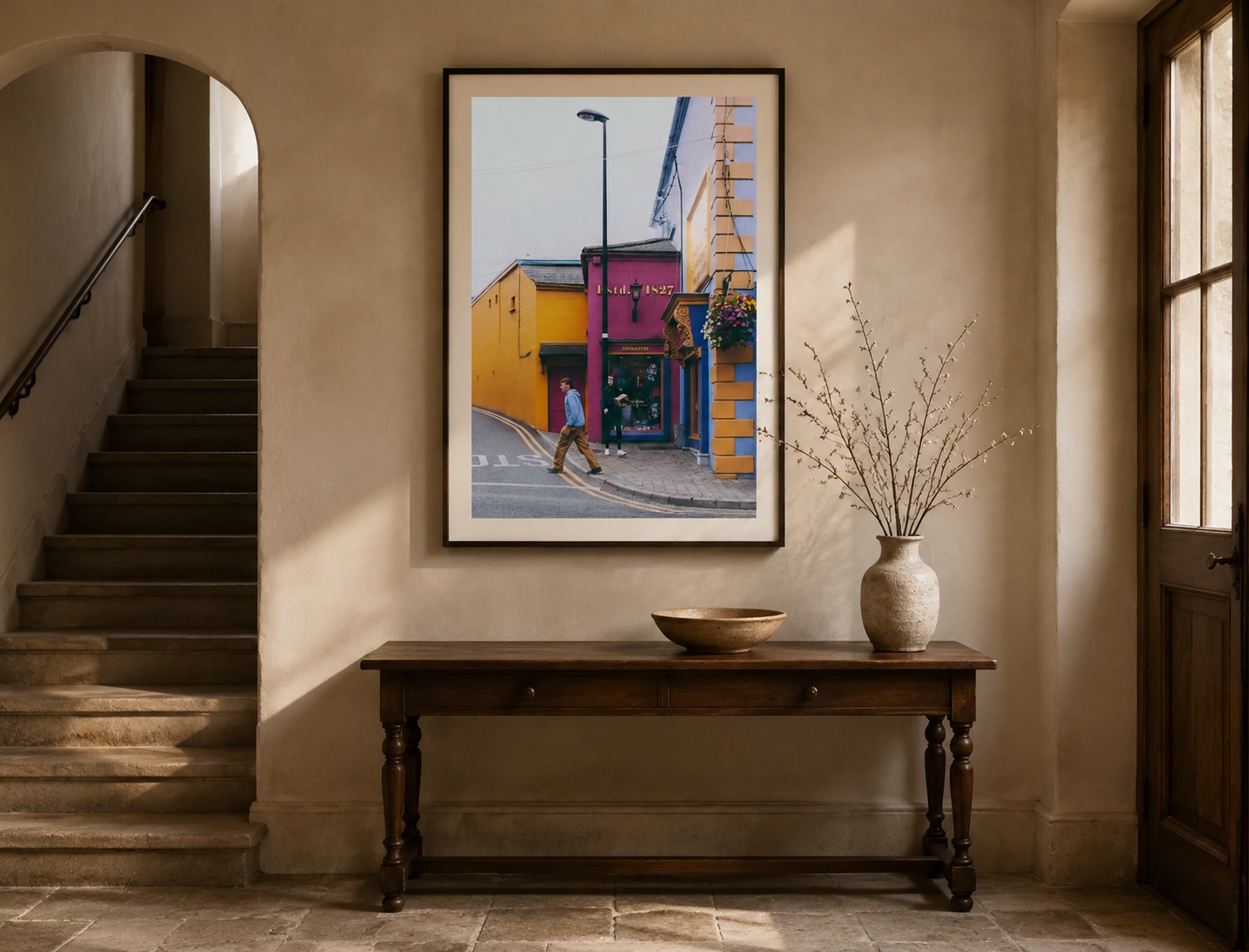 Interior of a house featuring a wooden console table with a large beige vase with bare branches and a shallow bowl. Behind the table is a framed colorful street scene with a person crossing a street, buildings painted in yellow, pink, and blue, and a streetlamp. To the left, stairs with a metal handrail lead up, and to the right is a window with wooden framing.