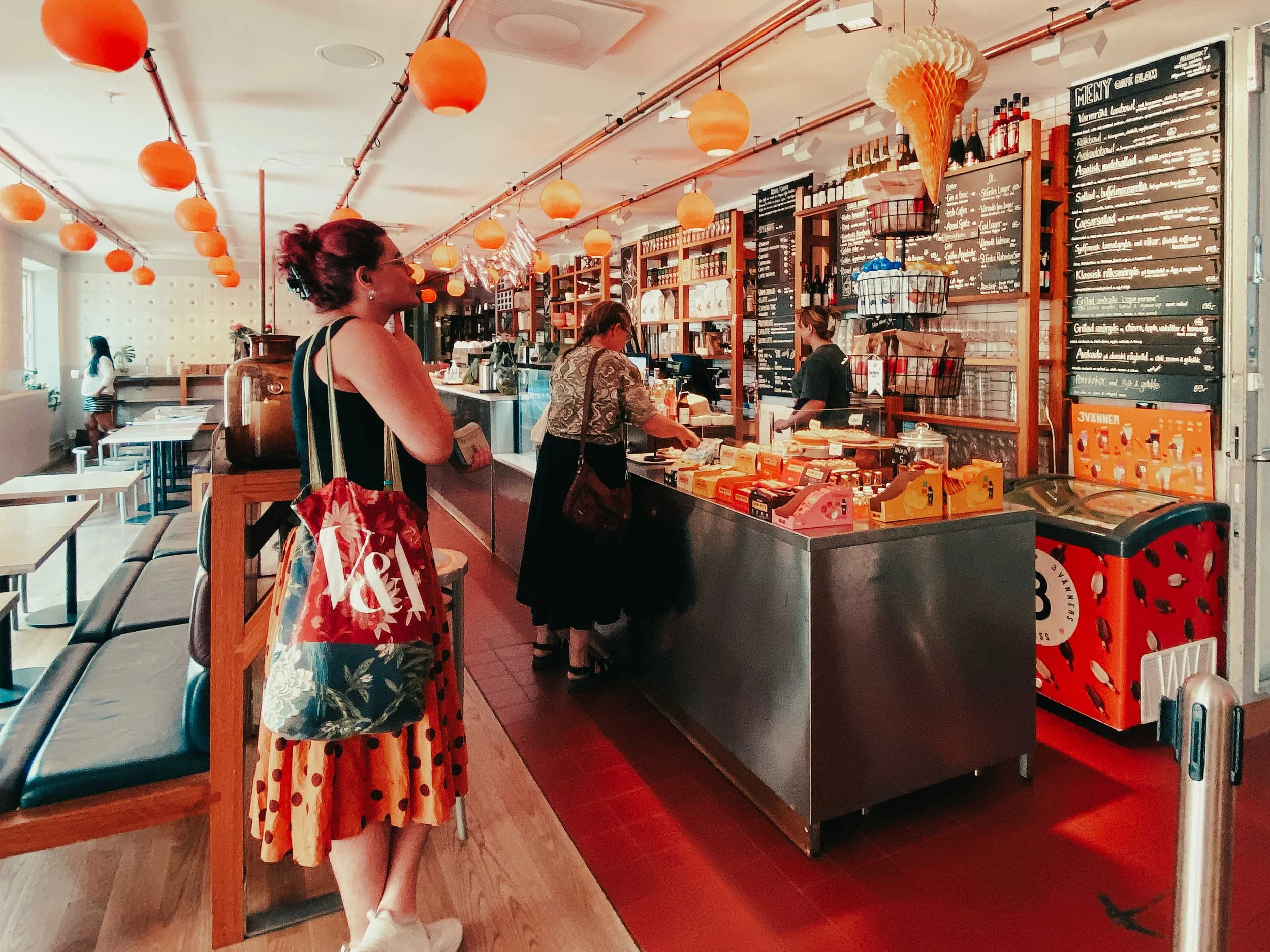 A woman in a polka dot orange skirt and floral blouse standing in line at a café counter, with others and menu boards in the background.