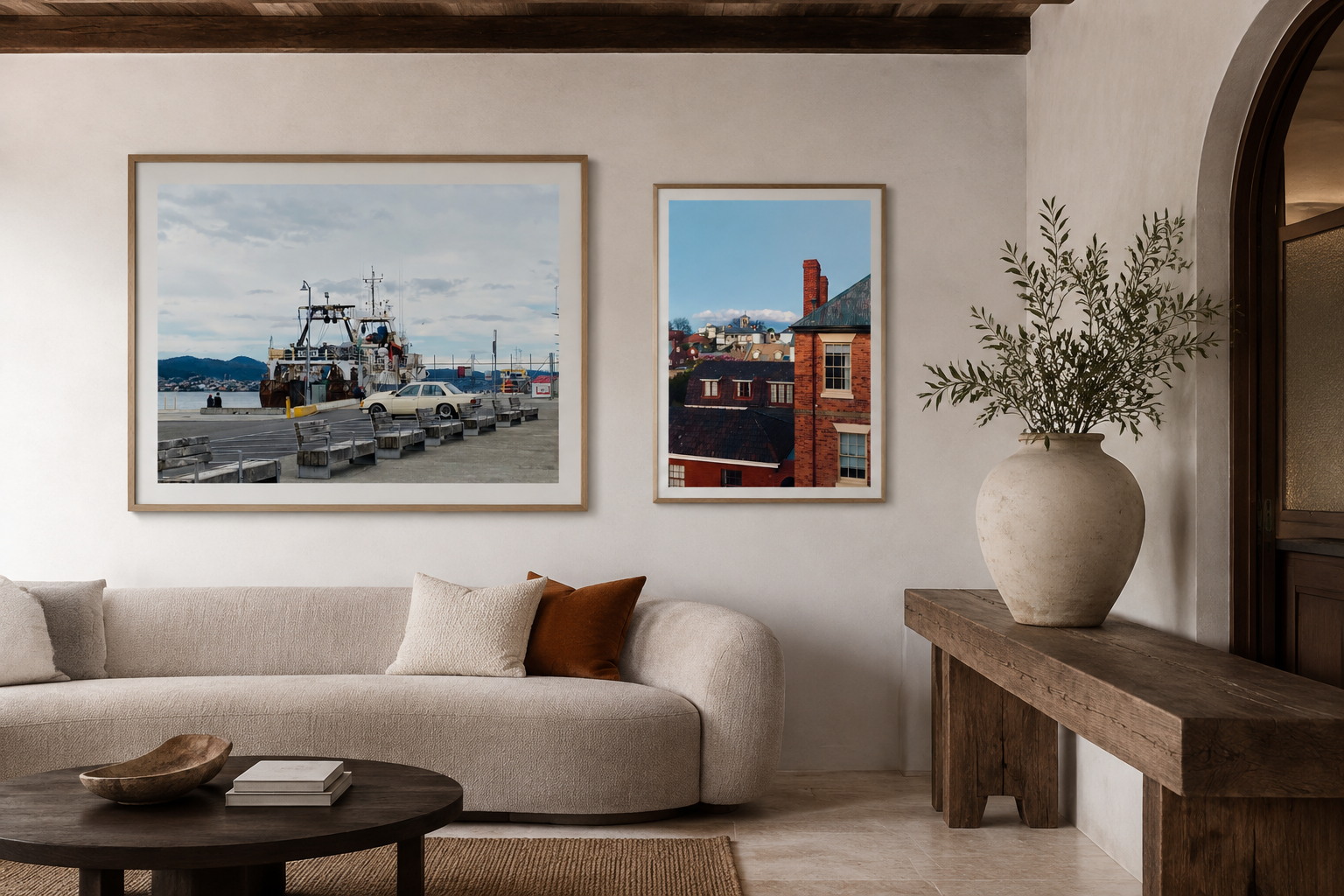 Living room with off-white sofa, beige and rust-colored pillows, wooden coffee table, large vase with greenery on side table, framed pictures of a harbor scene and city rooftops on the wall, beige walls, wooden ceiling beams, and an arched doorway.