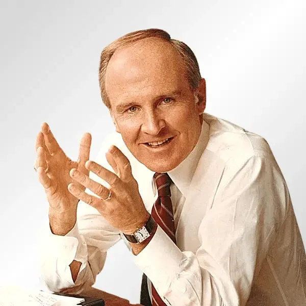 A middle-aged man with short light brown hair, wearing a white dress shirt, striped tie, and a watch, sitting at a desk with a cheerful expression, hands slightly raised as if in conversation, against a plain white background.