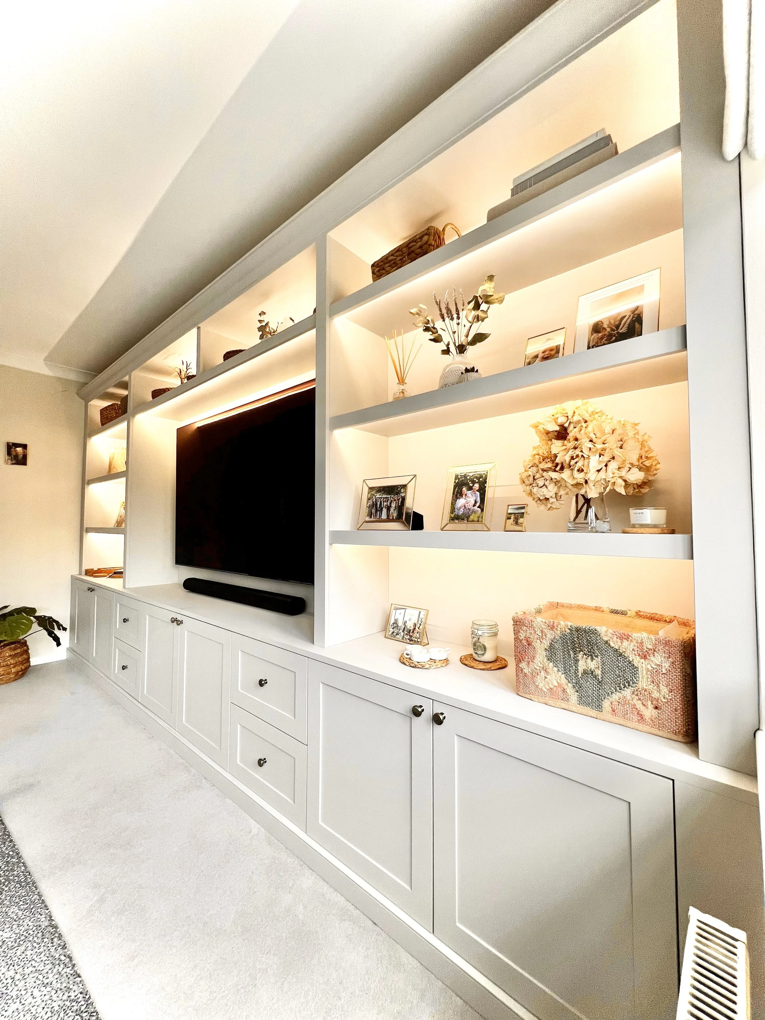 White built-in cabinet with drawers, shelves, framed photos, flowers in vases, and decorative items, above a white carpeted floor.