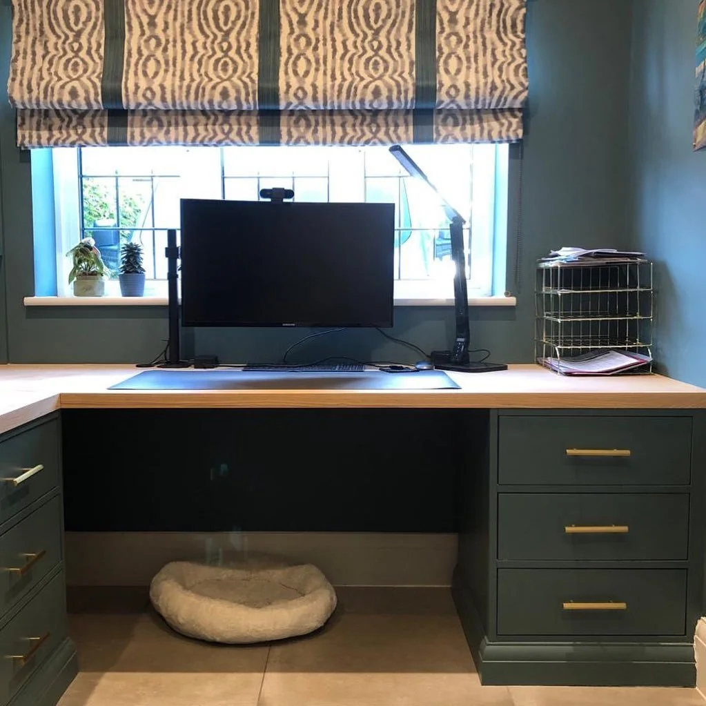 Home office workspace with a desk, a computer monitor, a desk lamp, a small shelf with papers, a window with patterned curtains, and a pet bed on the floor underneath the desk.