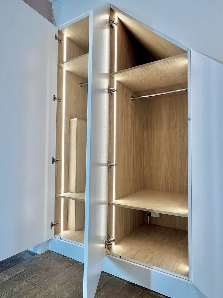 Open white wardrobe with multiple compartments and shelves, illuminated from the inside, on a wooden floor.