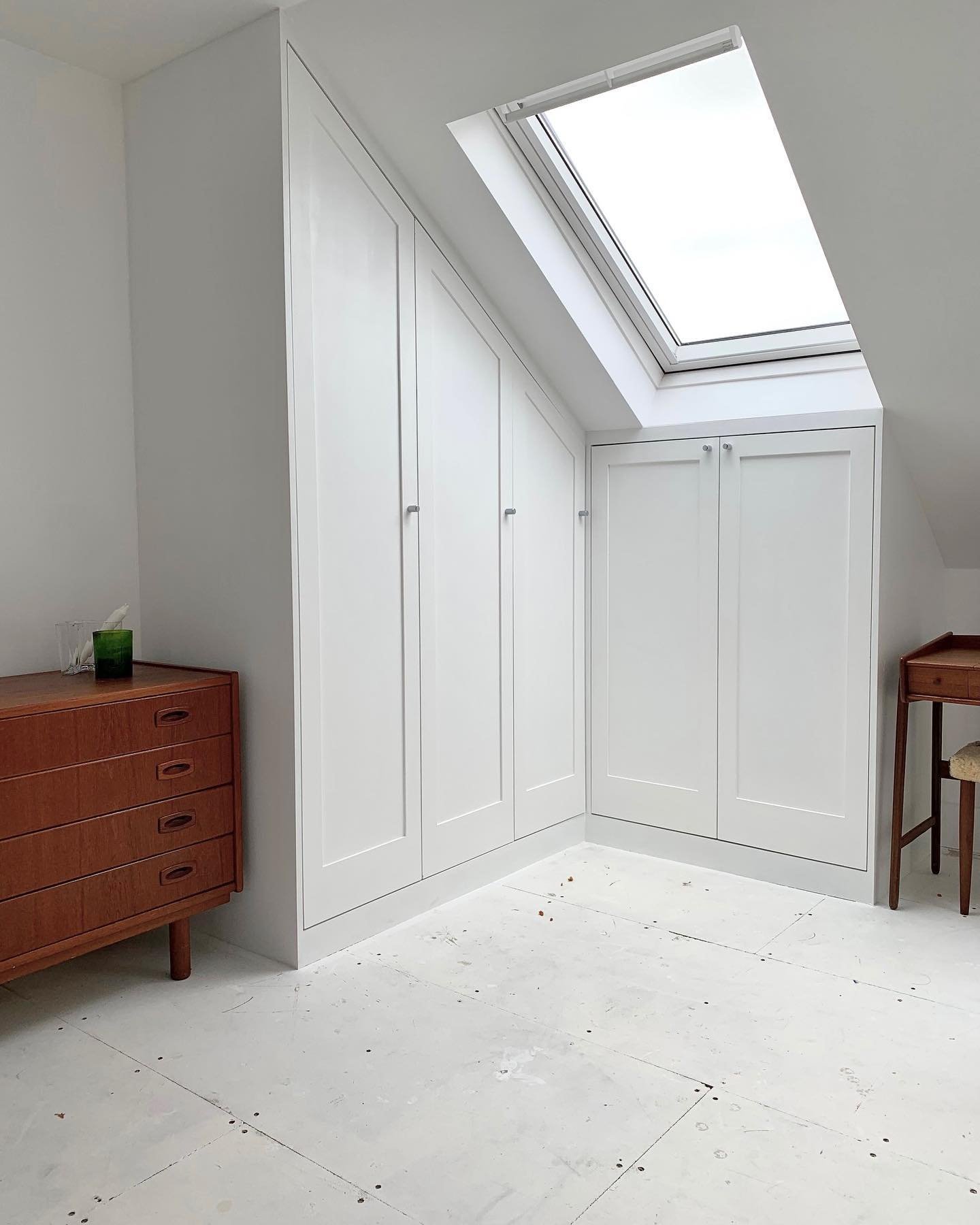 White built-in wardrobe with sloped ceiling and a skylight window, with two wooden dressers visible, one on the left and one on the right. The floor is white with some dirt or debris.