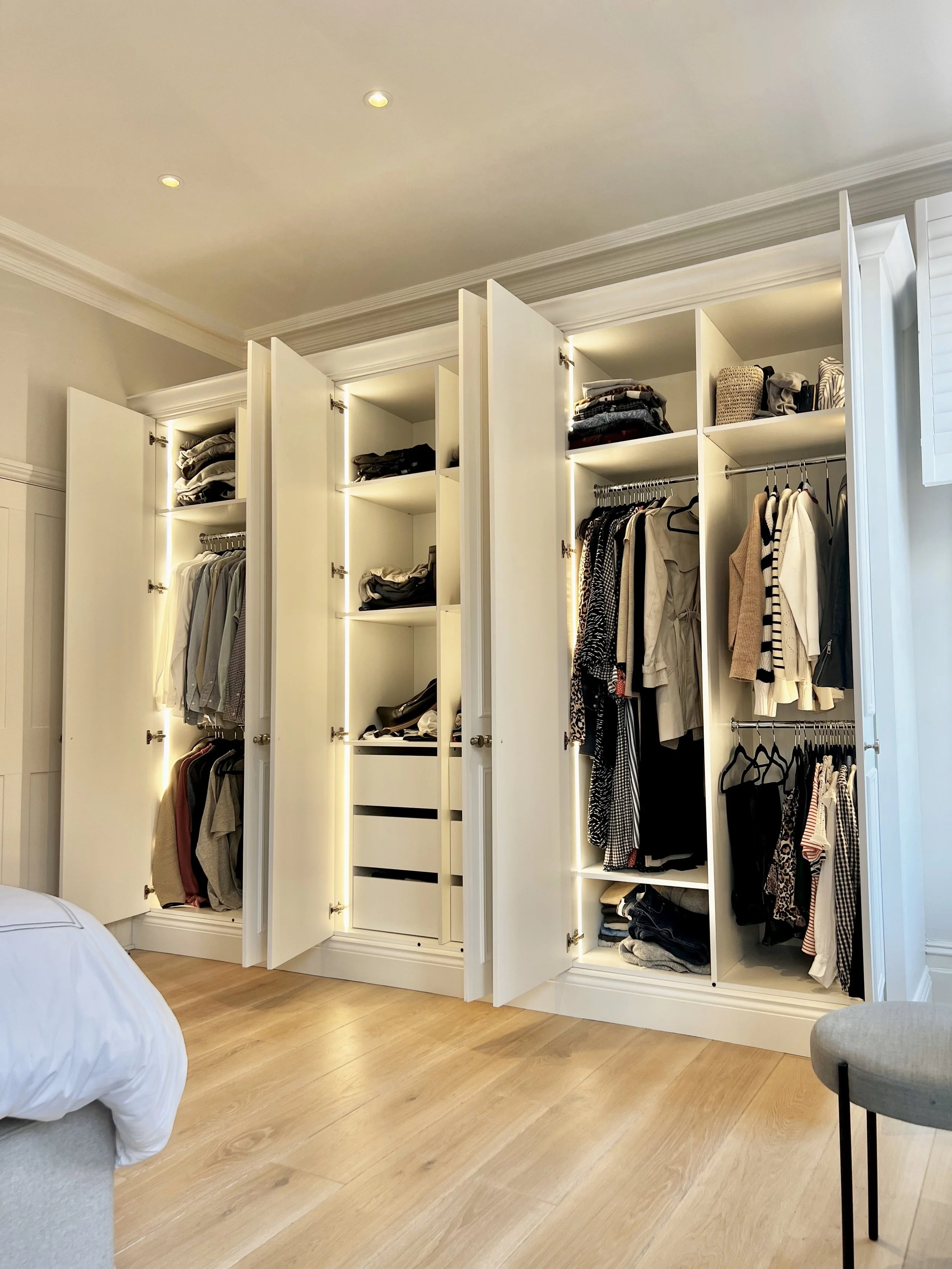 Open white wardrobe with multiple compartments filled with clothes, shoes, and accessories in a bright bedroom with wooden flooring.