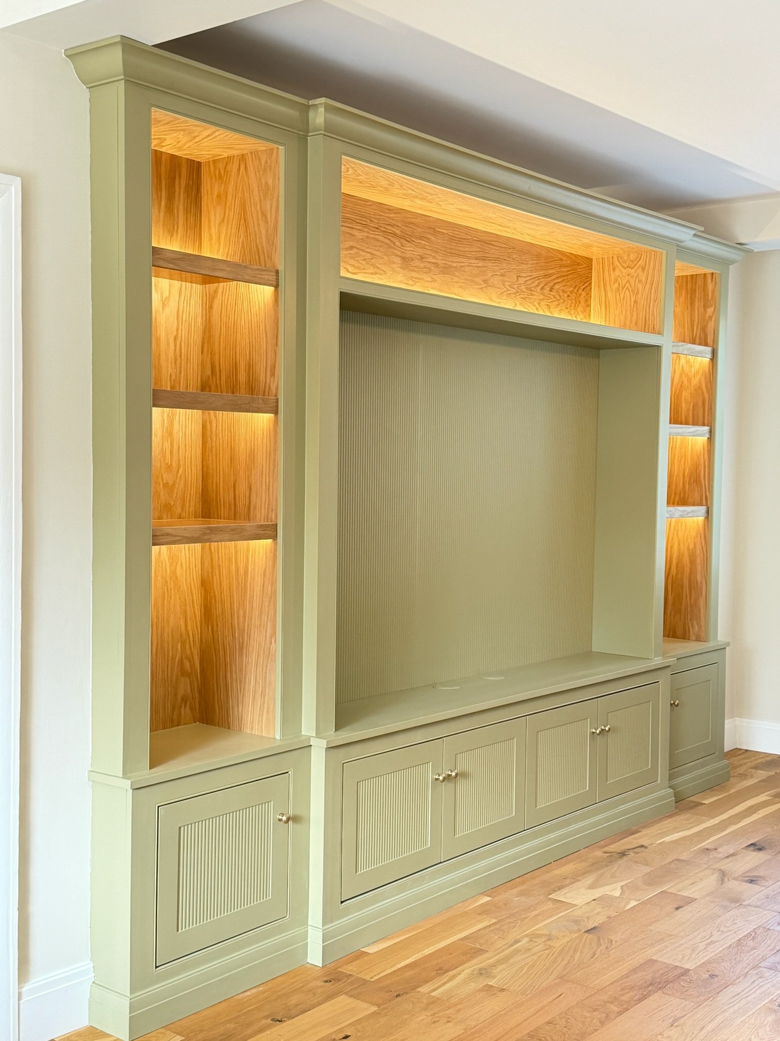 A built-in wall unit painted in light green with closed cabinets at the bottom, open shelves in the middle, and illuminated wooden shelves at the top, standing on a hardwood floor.