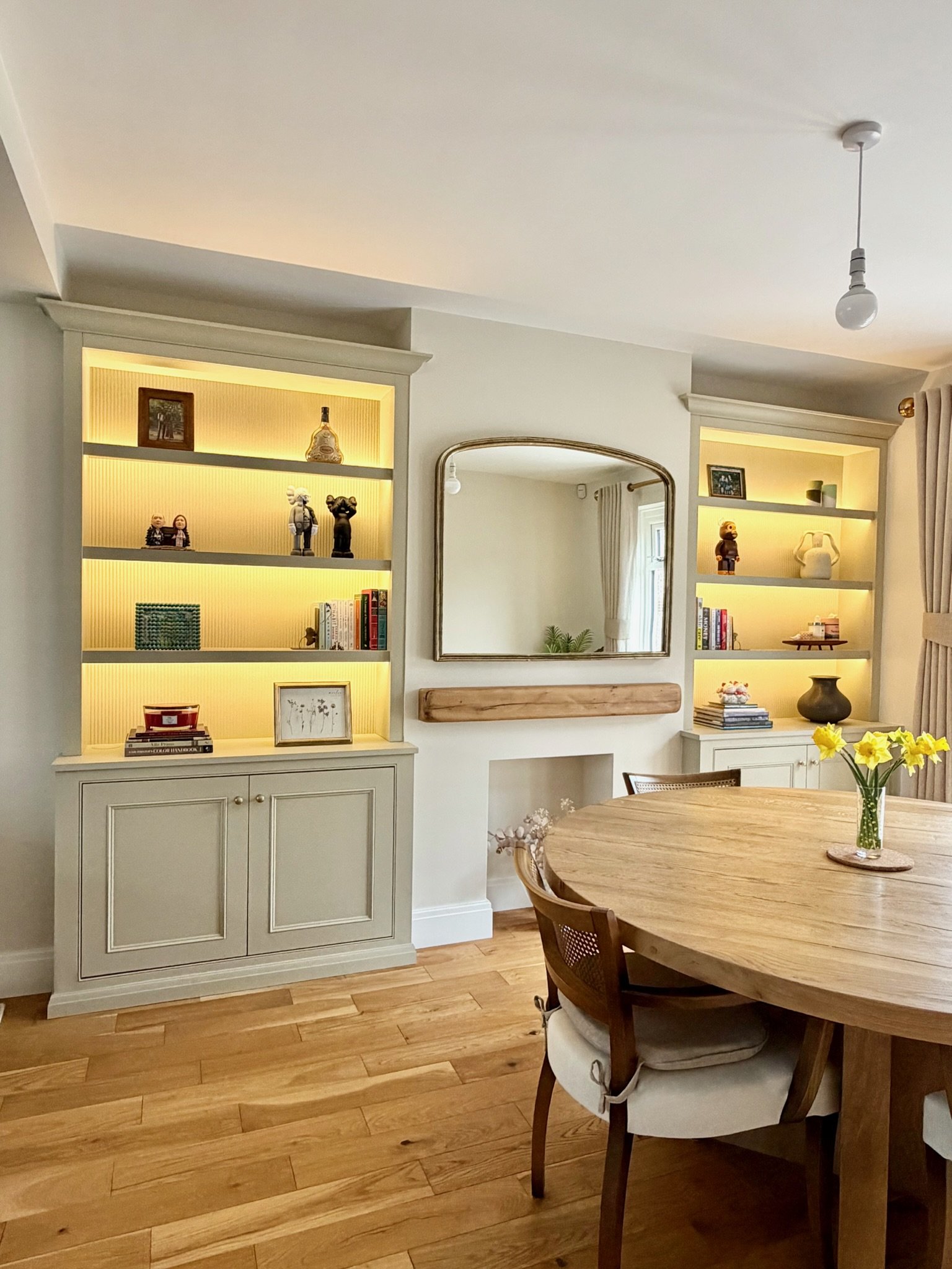 The image shows a cozy and well-lit dining and living area with light-colored walls and wooden flooring. There is a round wooden dining table with cushioned chairs, one of which is visible. A small vase with yellow flowers is on the table. Two tall, open bookshelves with backlighting flank a wall mirror. The shelves hold various decorative items, books, and framed photos. A curtain-adorned window is partially visible in the background.