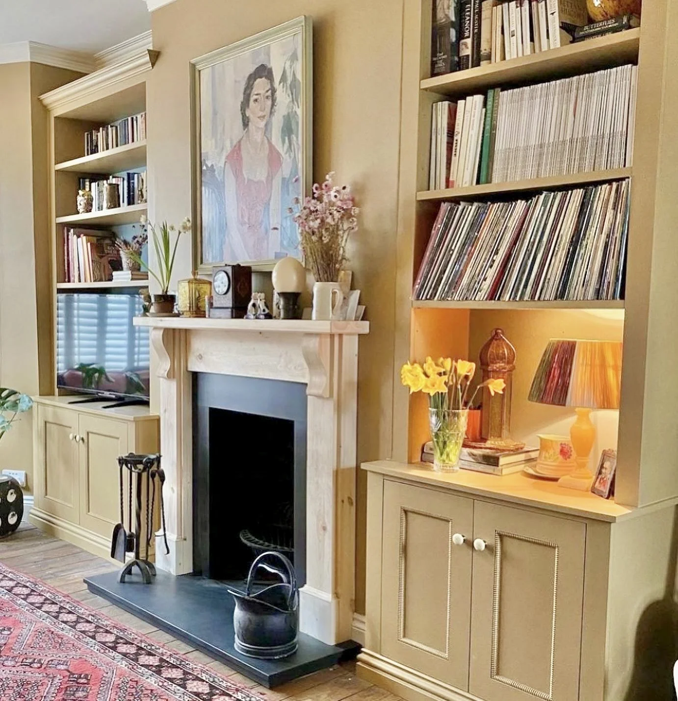 Living room corner with a fireplace, bookshelves, a flatscreen TV, and decorative items including flowers, a lamp, and framed photos.