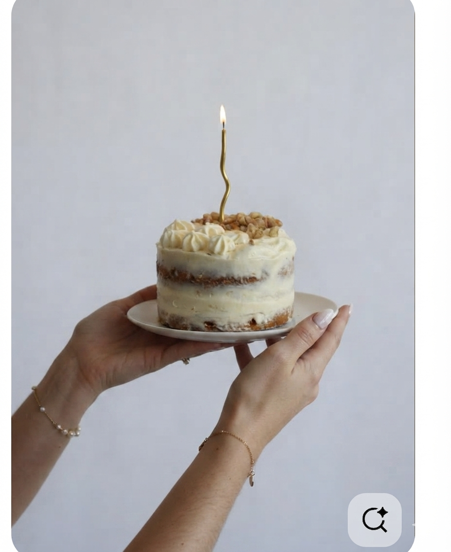 Person holding a small birthday cake with white frosting, topped with crushed nuts, and a lit gold birthday candle shaped like the number '1' against a plain light background.