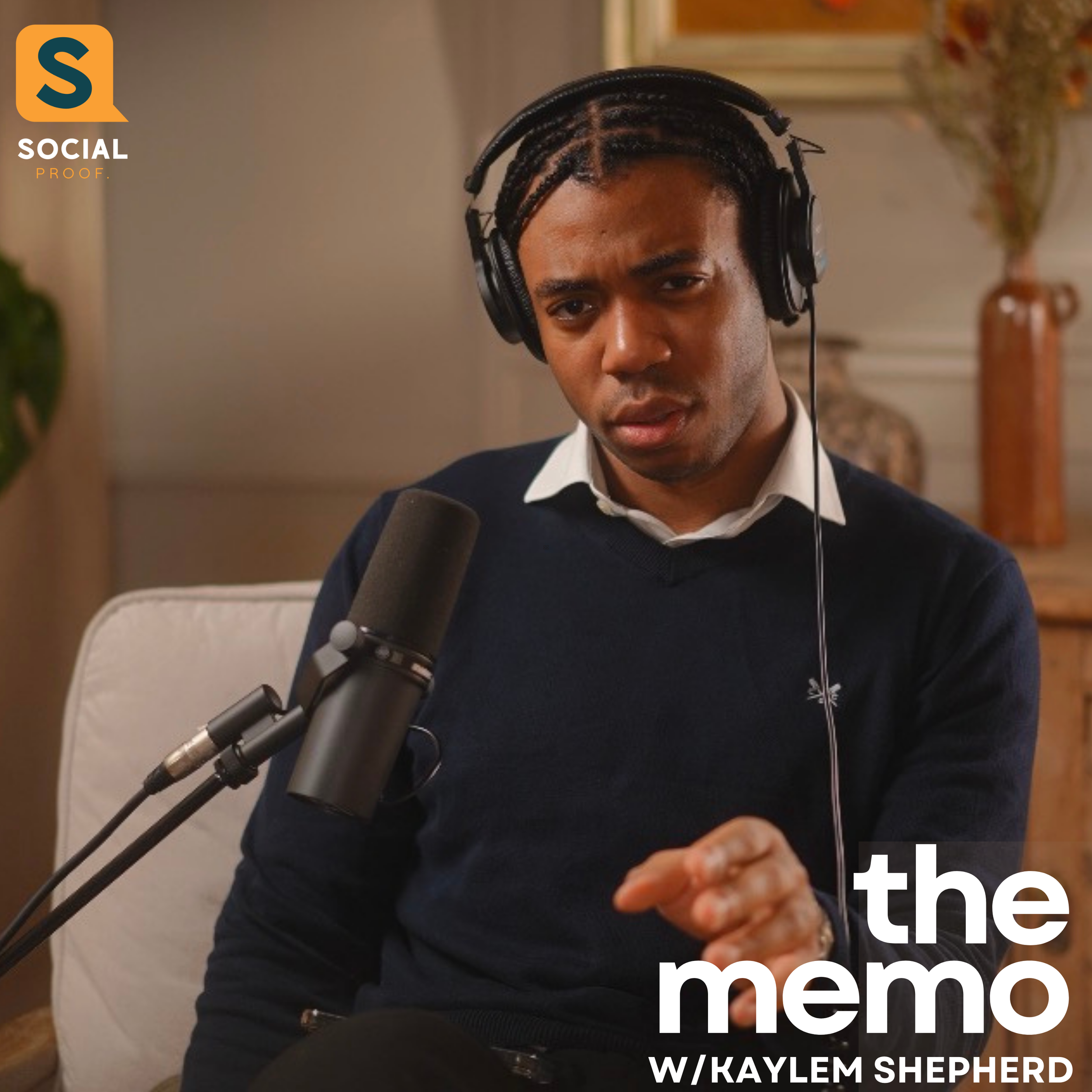 A man with braided hair and wearing a black sweater is speaking or singing into a microphone in an indoor setting, with a blurred background including a table with a vase and a framed picture.