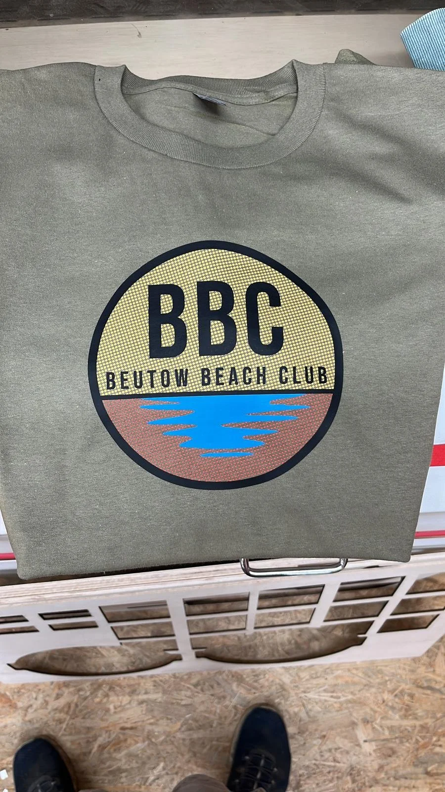 Ein graues T-Shirt mit einem runden Logo, das die Wörter 'BBC Beutow Beach Club' und eine stilisierte Wasserfläche zeigt.