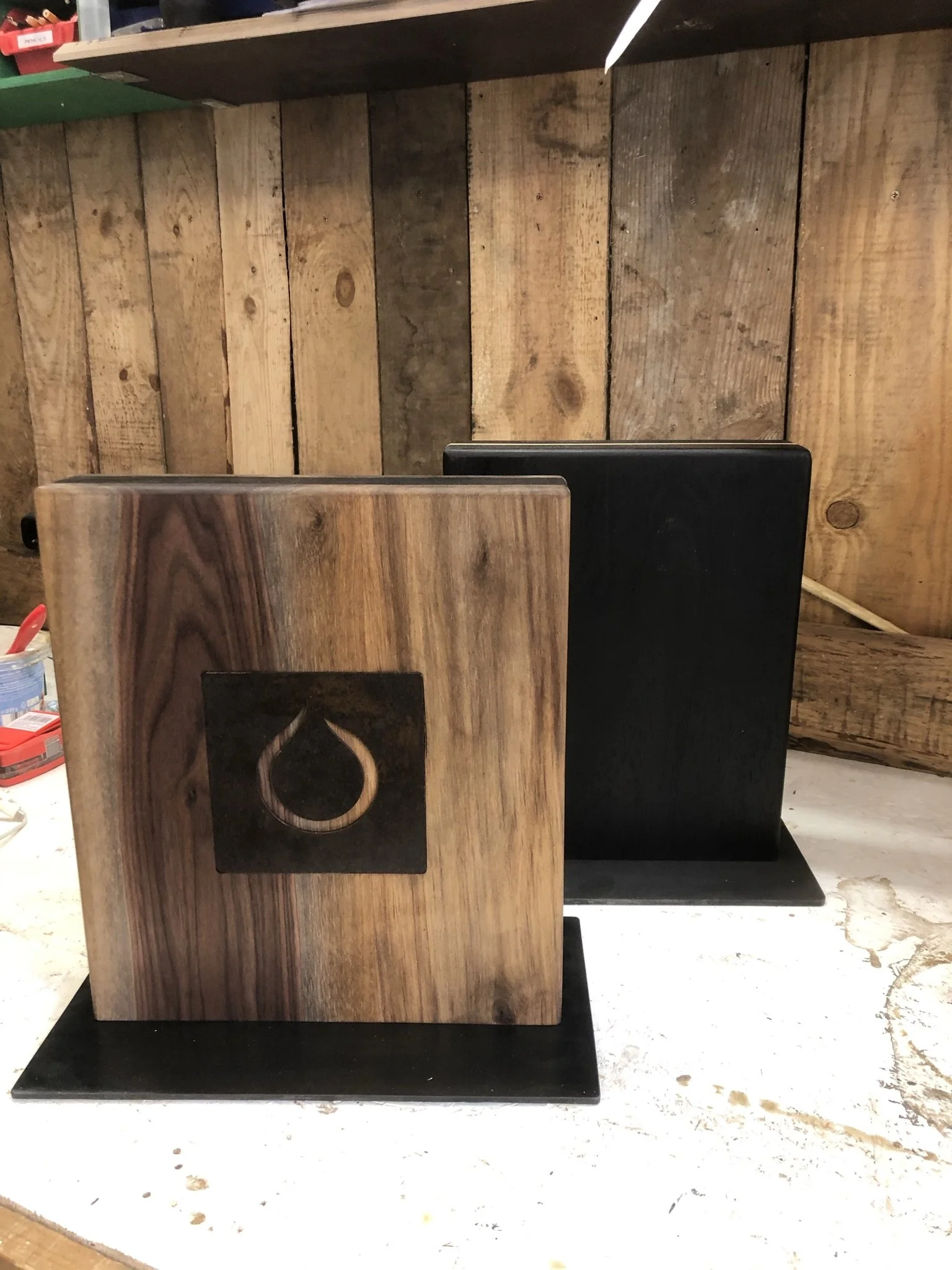 Zwei Holzboxen, eine helle mit dunklem Muster und eine schwarze, stehen auf einer schwarzen Unterlage auf einem Tisch vor einer Holzvertäfelung.