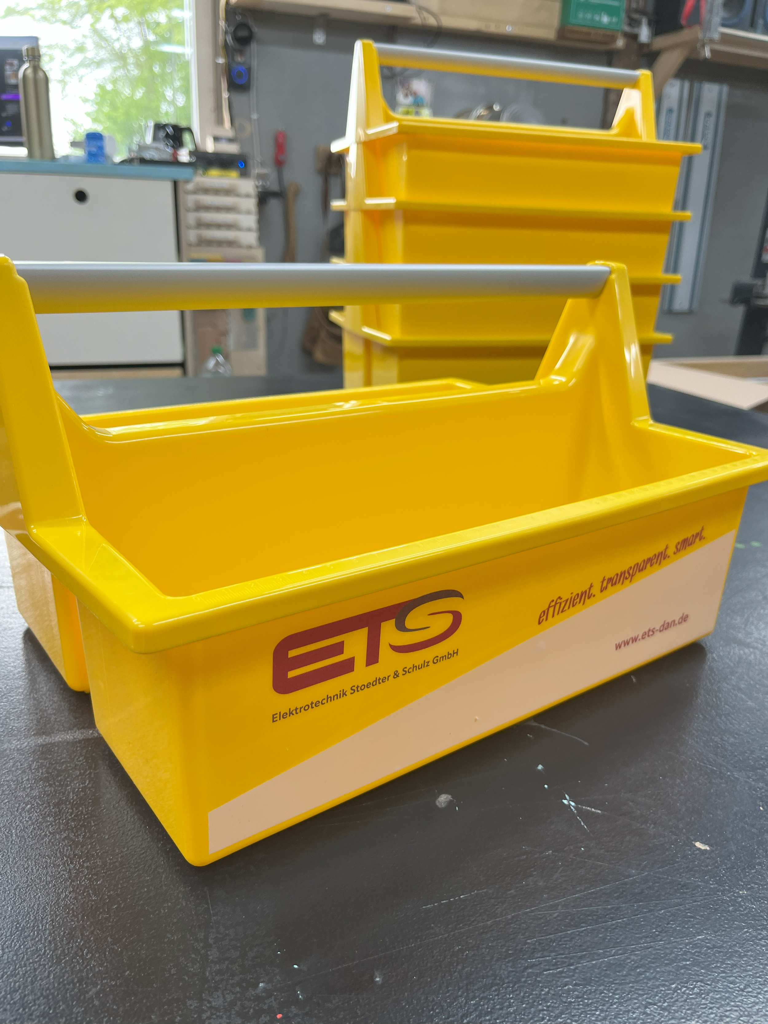 Gelbe Aufbewahrungsbox mit Logo und Text, steht auf einer schwarzen Oberfläche in einem Werkstatt- oder Lagerraum.