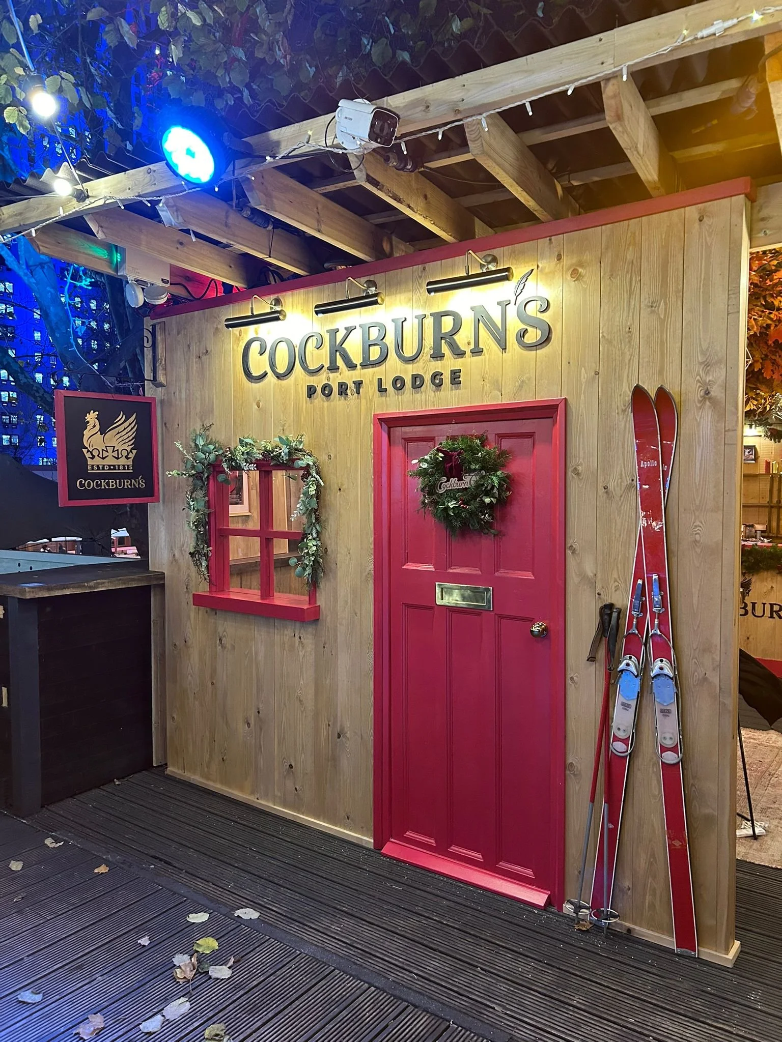 Holzständer mit rot lackiertem Türrahmen und Skiern, Weihnachtskränze und eine leuchtende Cockburns-Beschilderung bei Nacht.