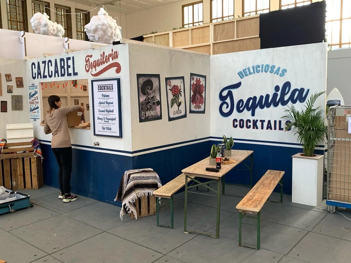Stand mit Tequila Cocktails in einer Halle, dekori‌ert mit Wolken und Gemälden, Tischen und Bänken, Person bestellt an einer Theke.