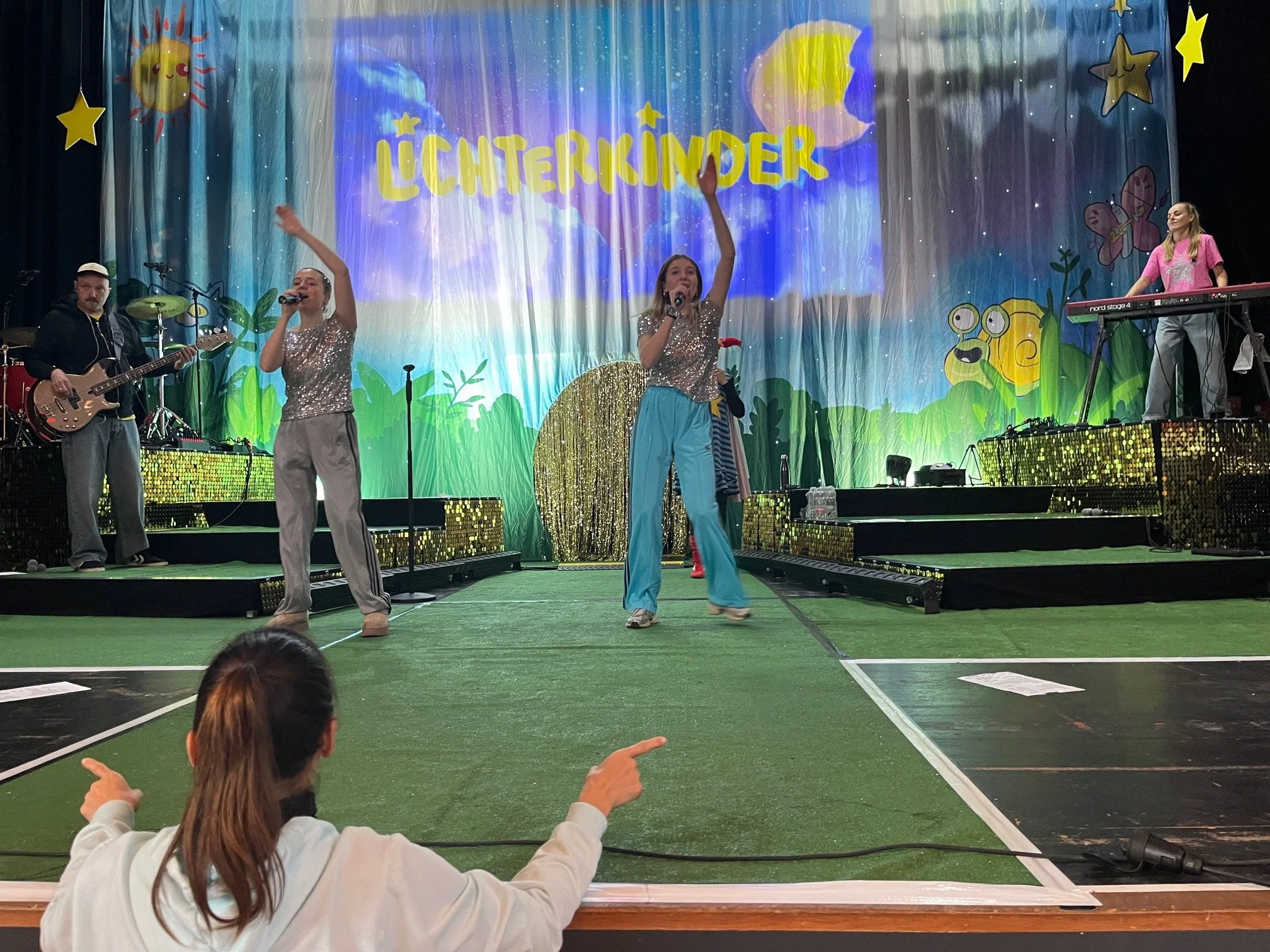 Auf der Bühne performen Kinder mit Musikern vor einem bunten Himmels-Hintergrund mit der Aufschrift 'Lichterkinder'.