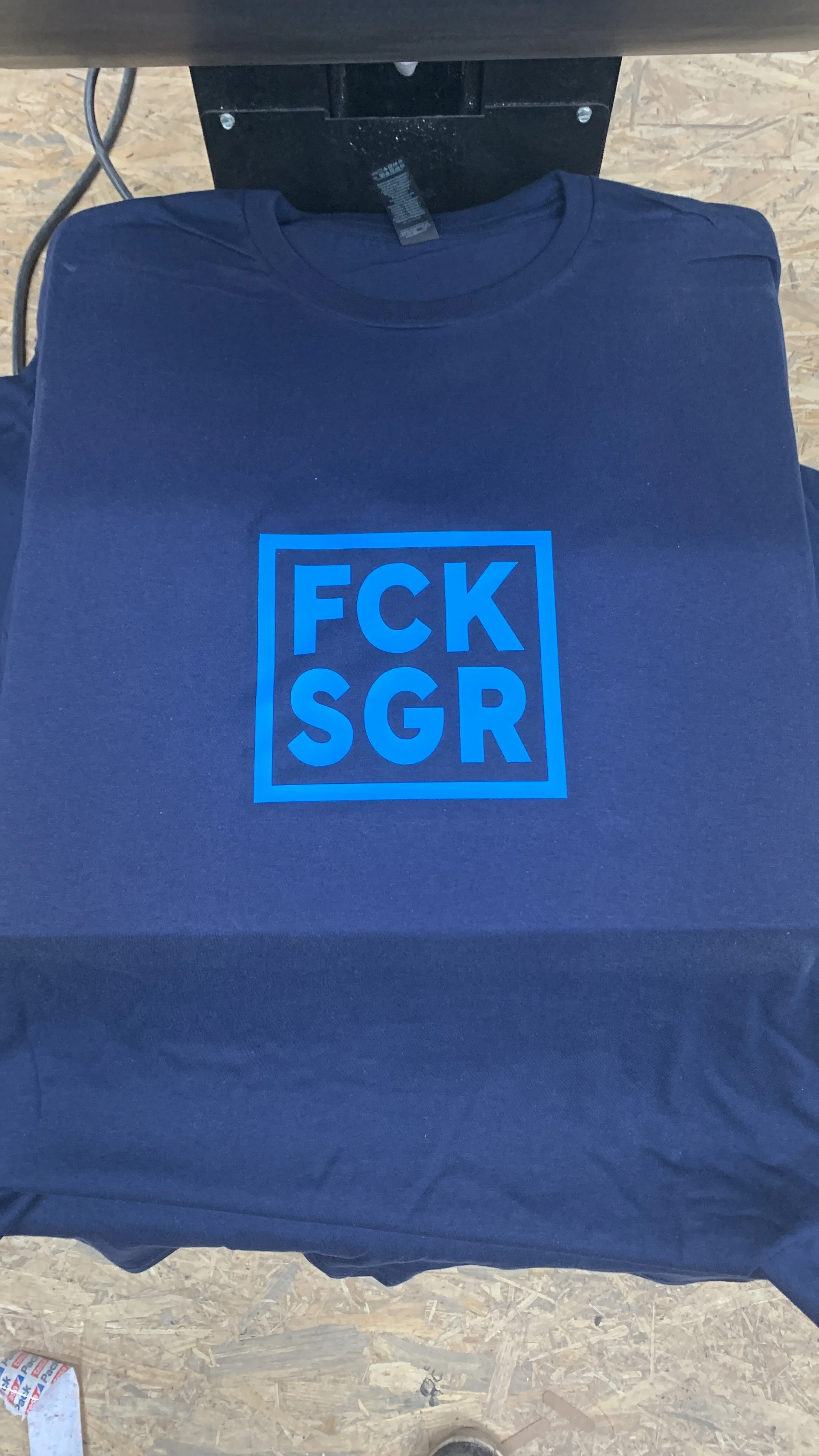 Ein schwarzes T-Shirt mit blauer Schrift in einem rechteckigen Rahmen, das die Worte 'FCK SGR' zeigt, liegt auf einem Tisch.