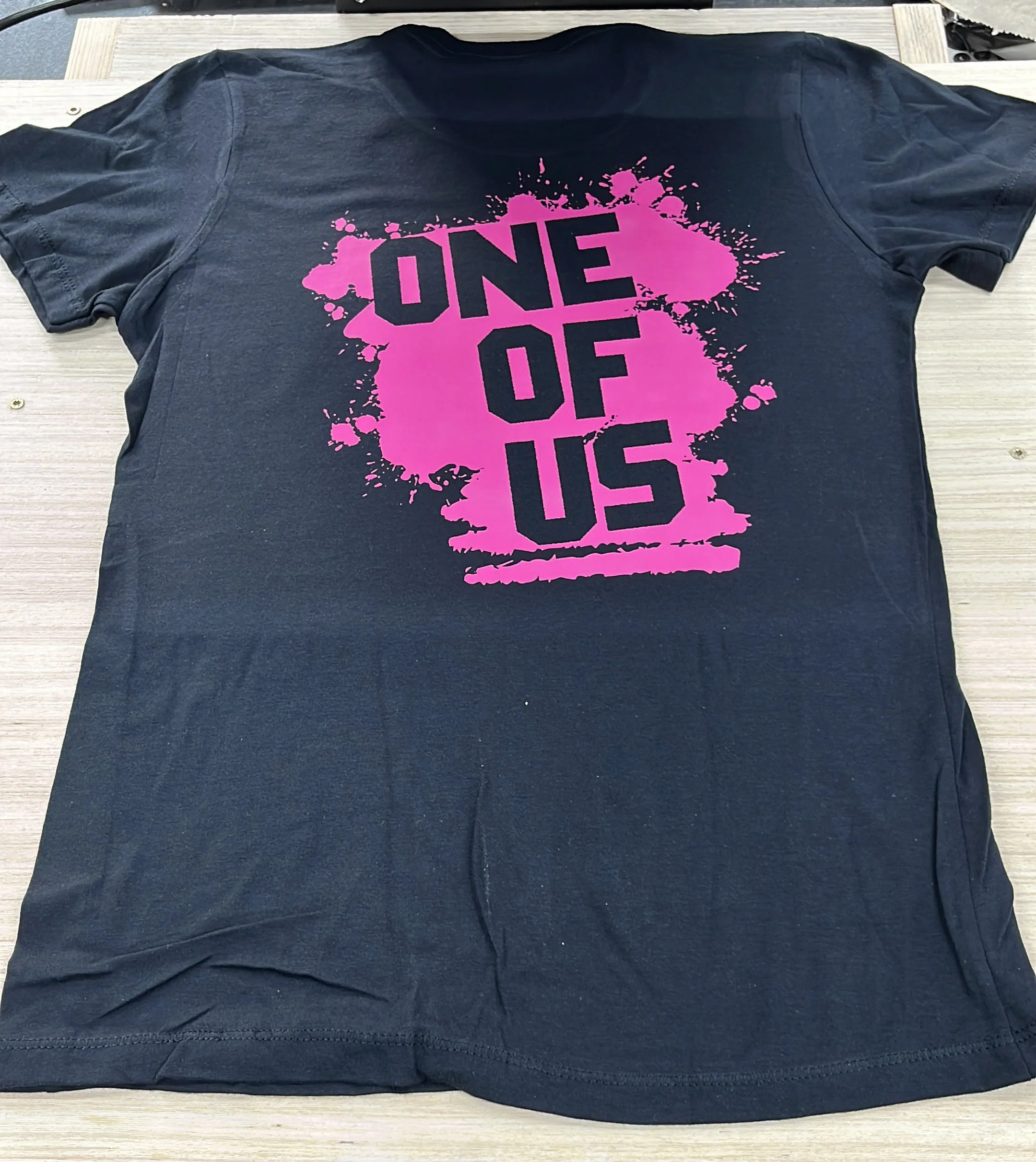 Schwarzes T-Shirt mit pinkem Spritzguss-Design mit der Aufschrift 'ONE OF US' auf dem Rücken, liegend auf einem Holztisch.