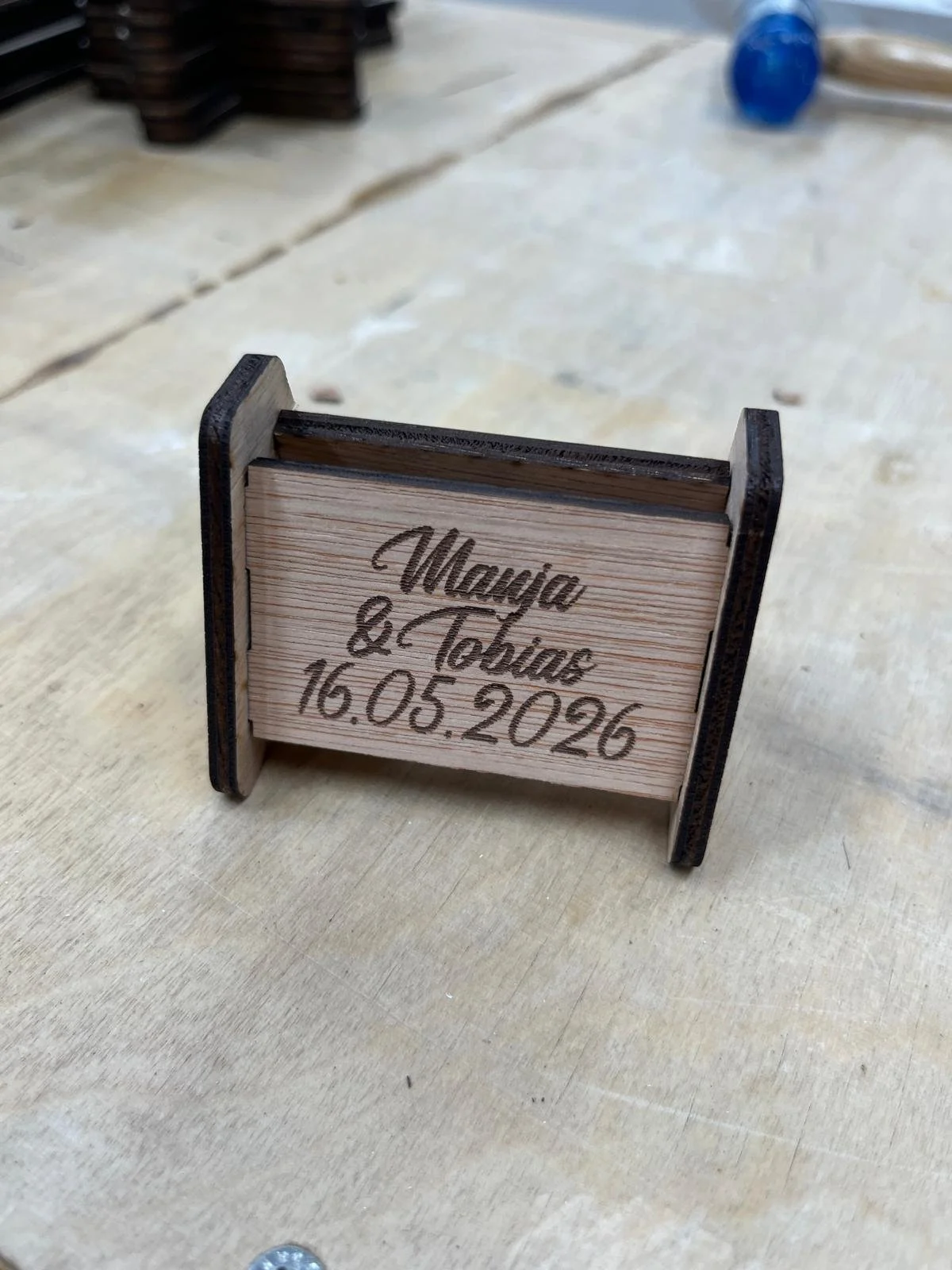 Ein kleines Holzschild mit graviertem Text über die Namen Manya und Tobias und das Datum 16.05.2026. Das Schild ist auf einem Tisch.