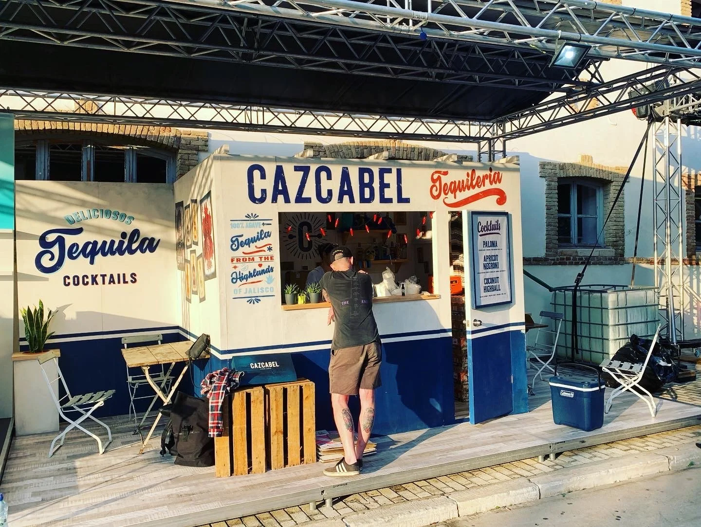 Ein stand namens 'CAZCABEL Tequileria' verkauft Cocktails, darunter Paloma, Apricot Negroni und Kokosnuss-Hibiskus. Es gibt einen Sitzbereich mit Stühlen und Tischen, eine Kühlbox, und eine Person bestellt einen Cocktail am Fenster. Der Stand ist im 