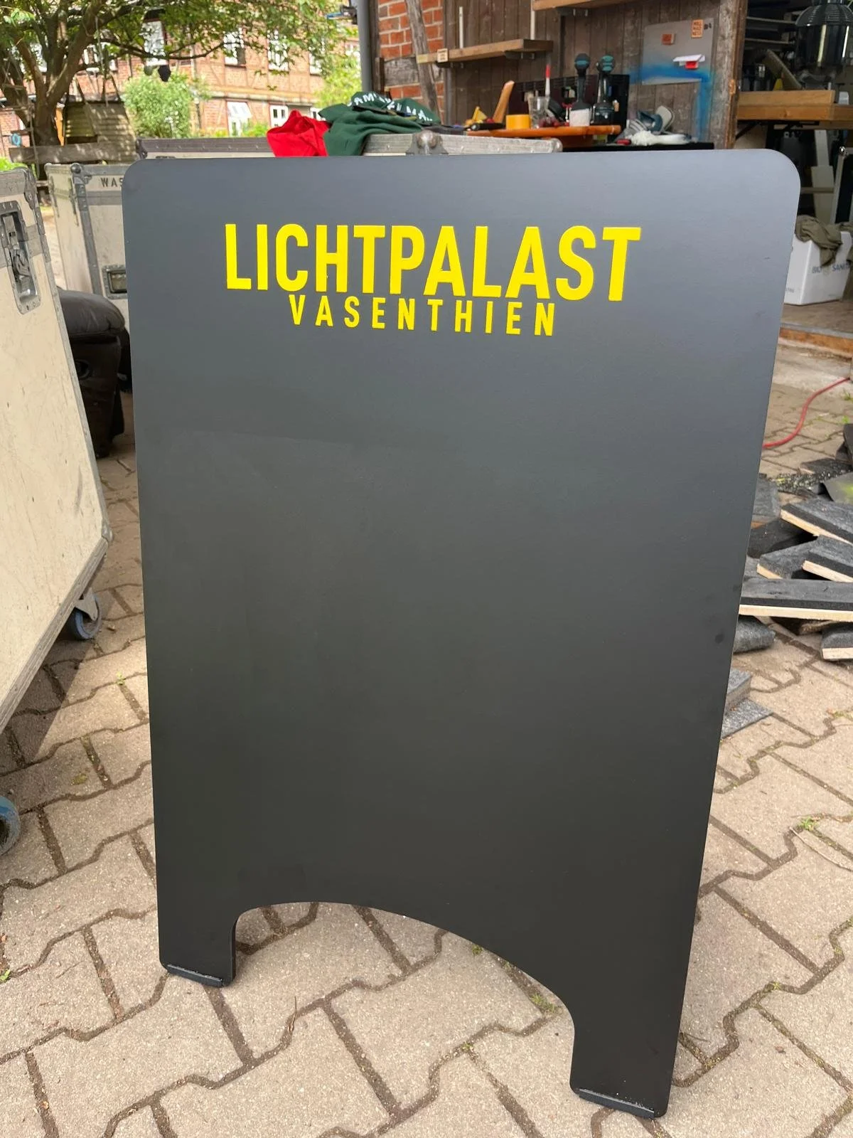 Schwarze Tafel mit gelben Buchstaben, die 'LICHTPALAST VASENTHIEN' sagen, auf einem Gehweg im Freien, im Hintergrund Werkzeuge und Arbeitsmaterialien