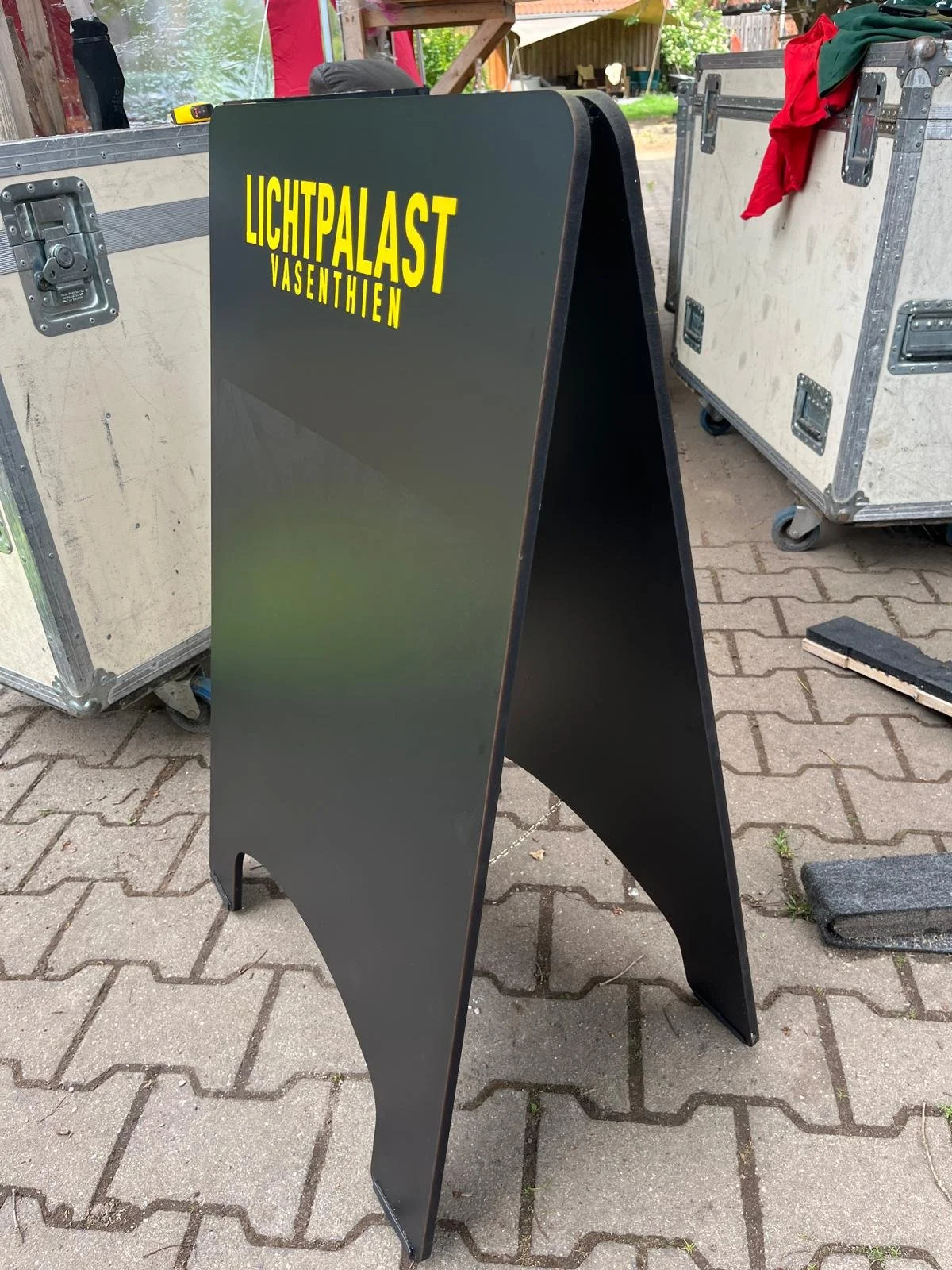 Ein schwarzer, schulterhoher Pult mit gelber Aufschrift »LICHTPALAST VASENTHIEN« auf einer Gehwegplatte, umgeben von Umzugskartons und anderen Gegenständen im Freien.